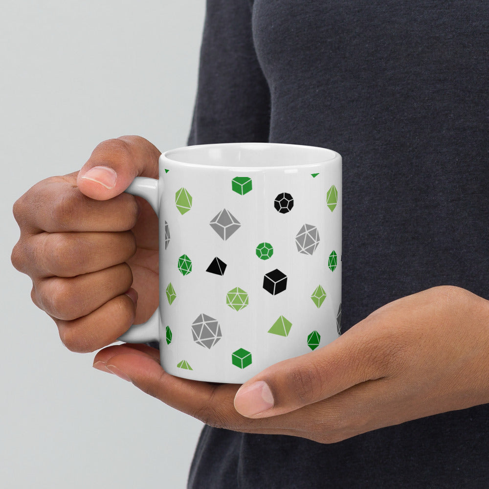 white mug being held by the handle by a dark-skinned set of hands. It has an all-over print of polyhedral d&d dice in the aromantic colors of greens, grey, and black