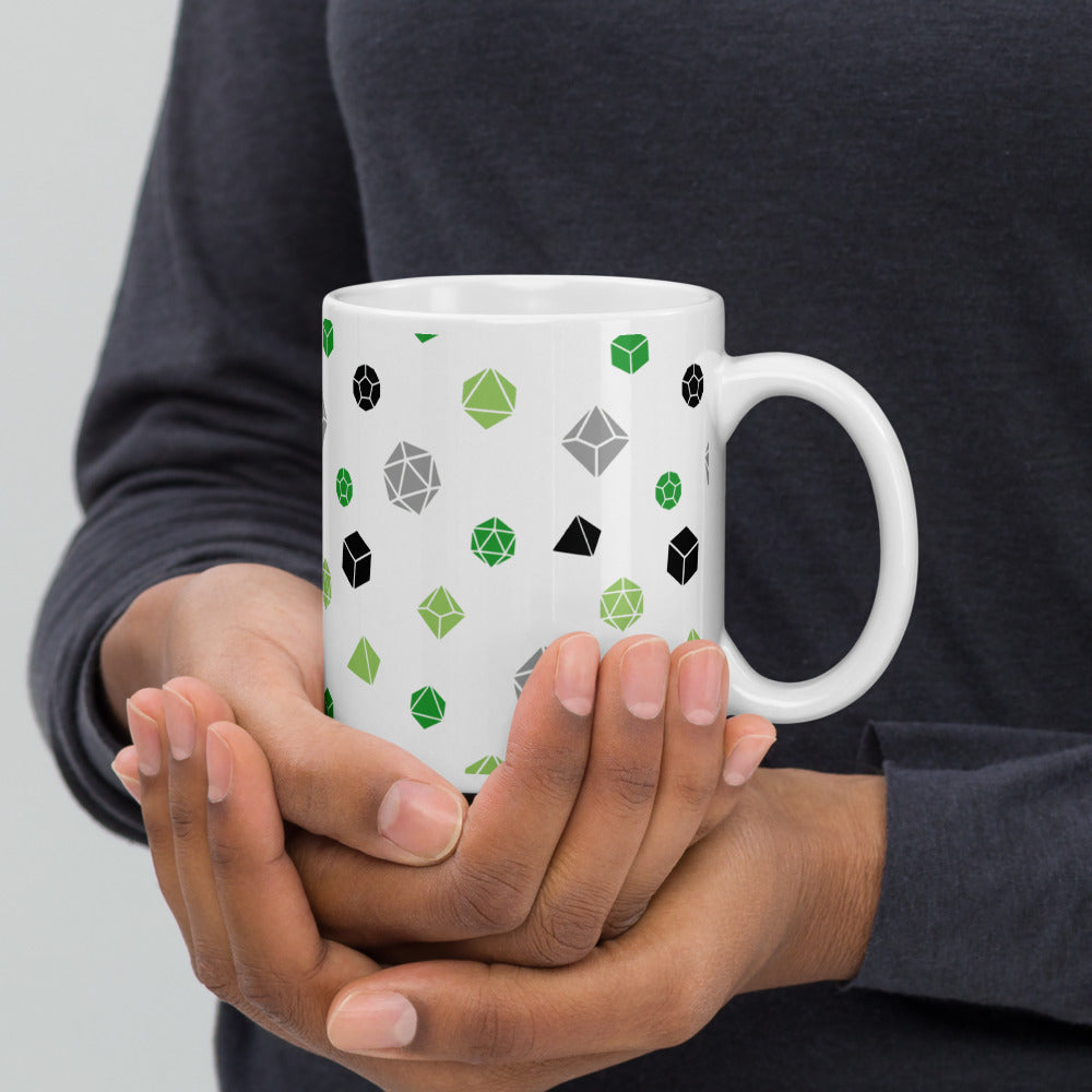 white mug being cupped by a dark-skinned set of hands. It has an all-over print of polyhedral d&d dice in the aromantic colors of greens, grey, and black