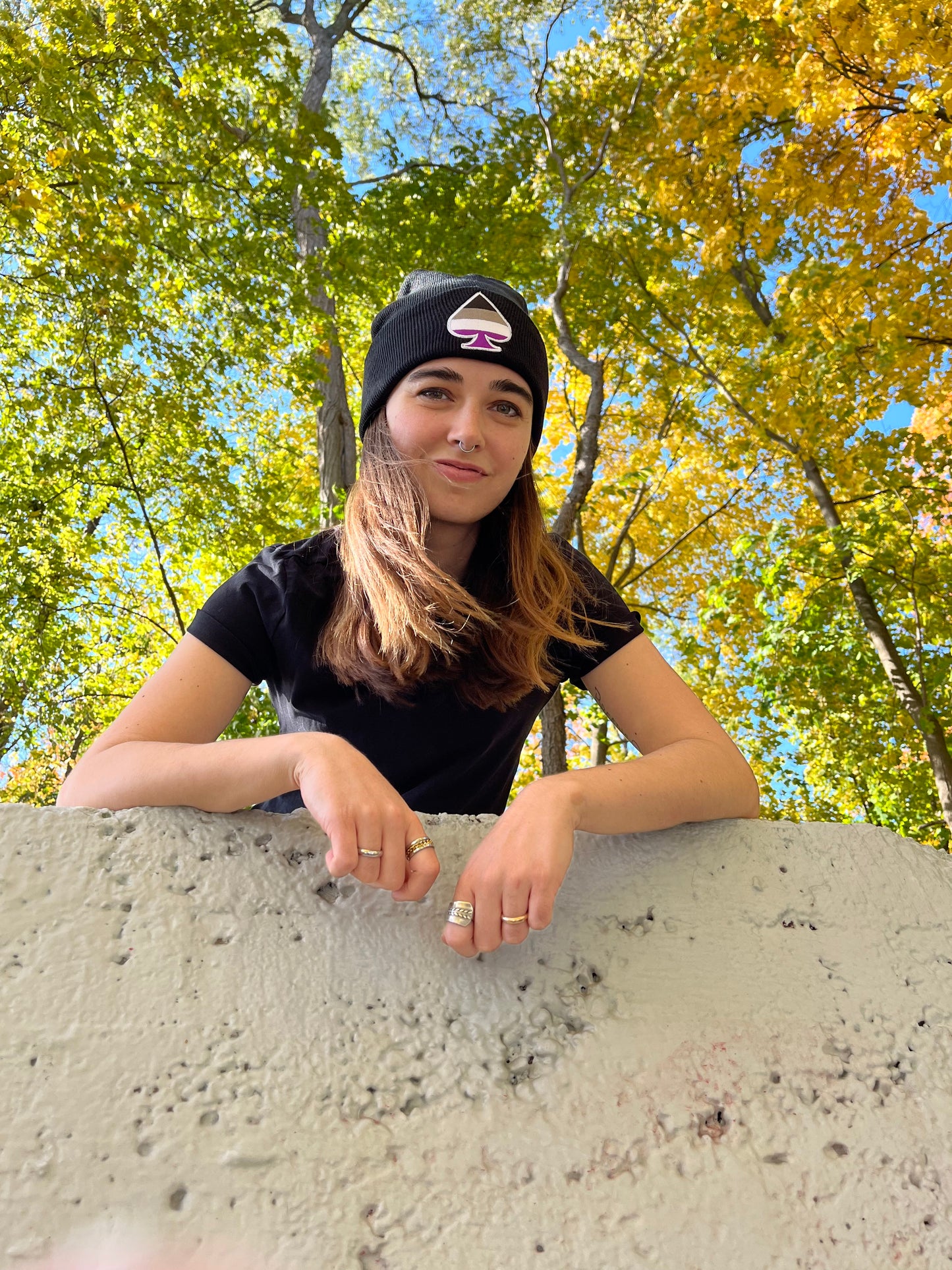 waist up shot of smiling person leaning against white wall with trees behind and wearing black asexual pride beanie