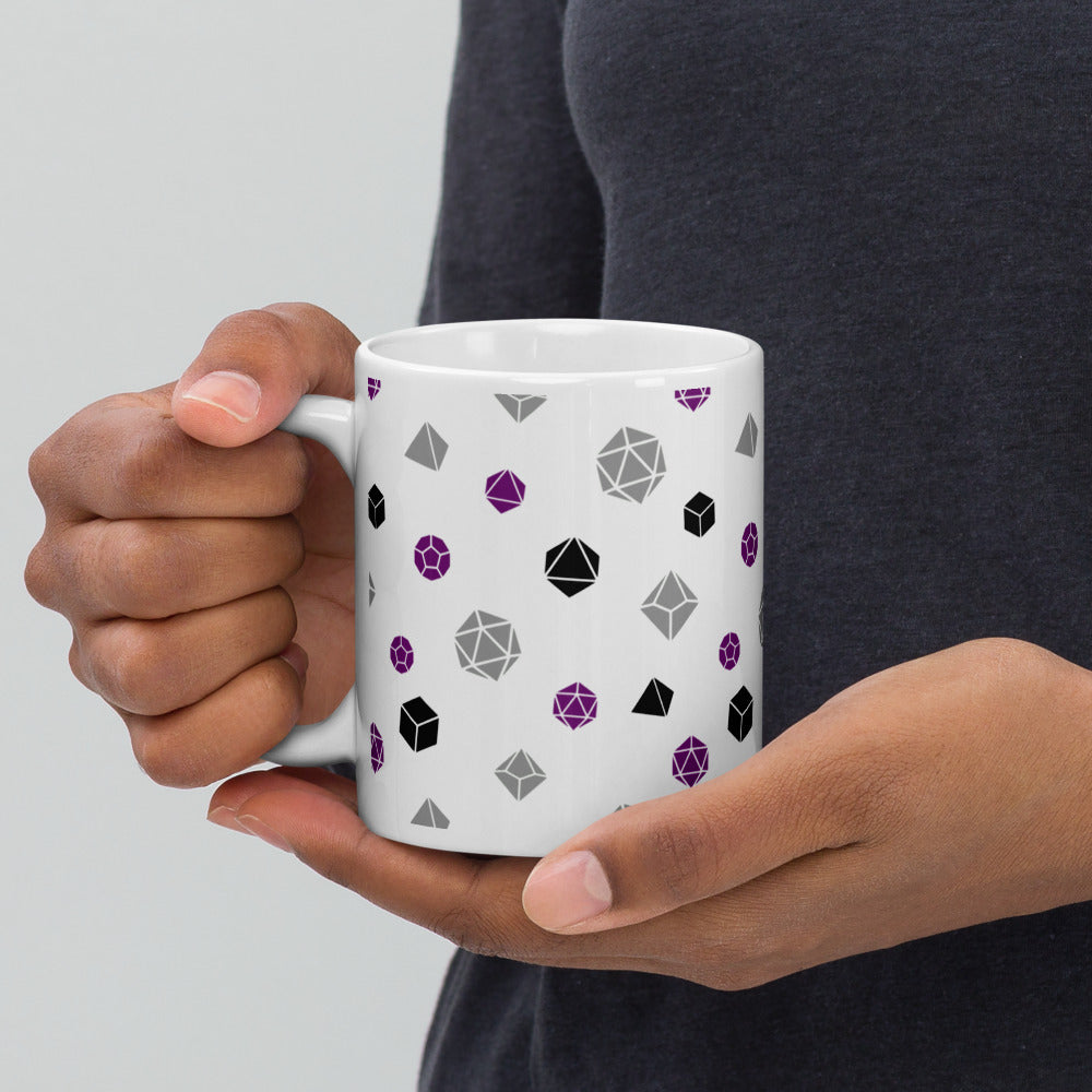 white mug being held by the handle by a dark-skinned set of hands. It has an all-over print of polyhedral d&d dice in the asexual colors of purple, grey, and black
