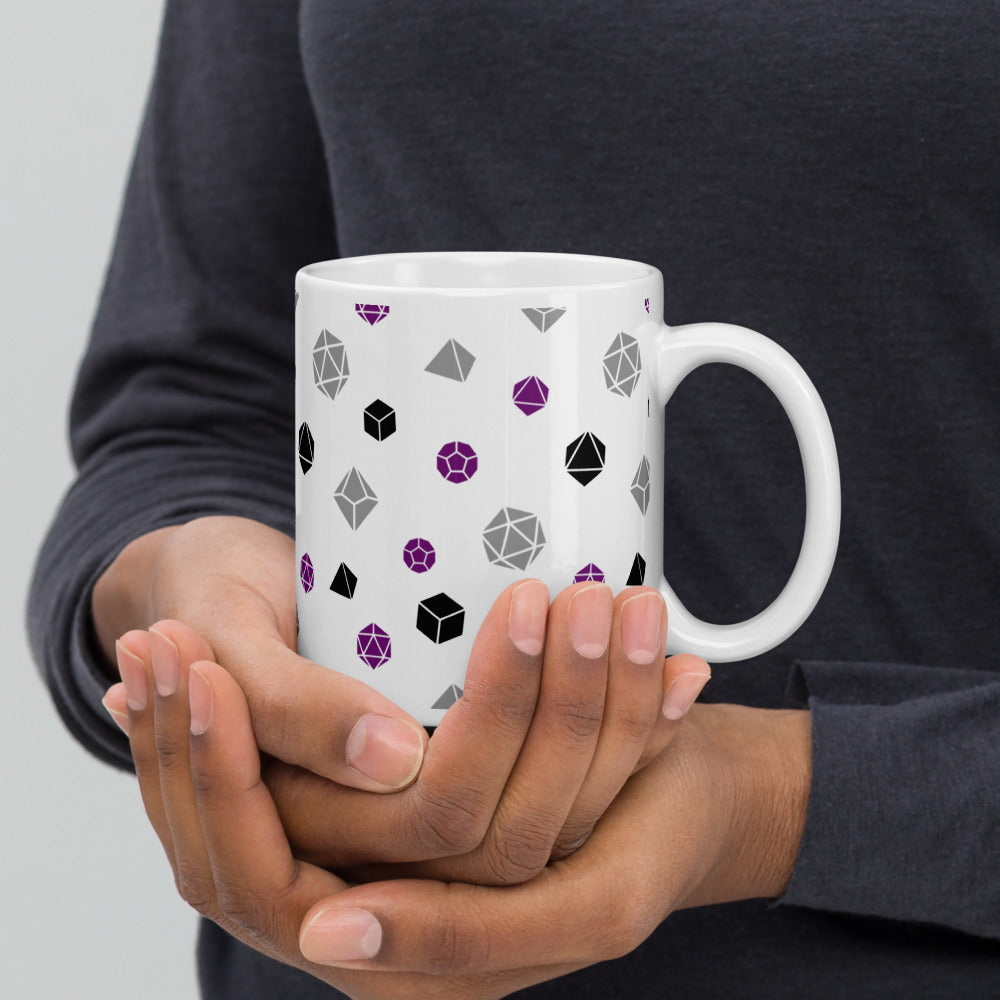 white mug being cupped by a dark-skinned set of hands. It has an all-over print of polyhedral d&d dice in the asexual colors of purple, grey, and black
