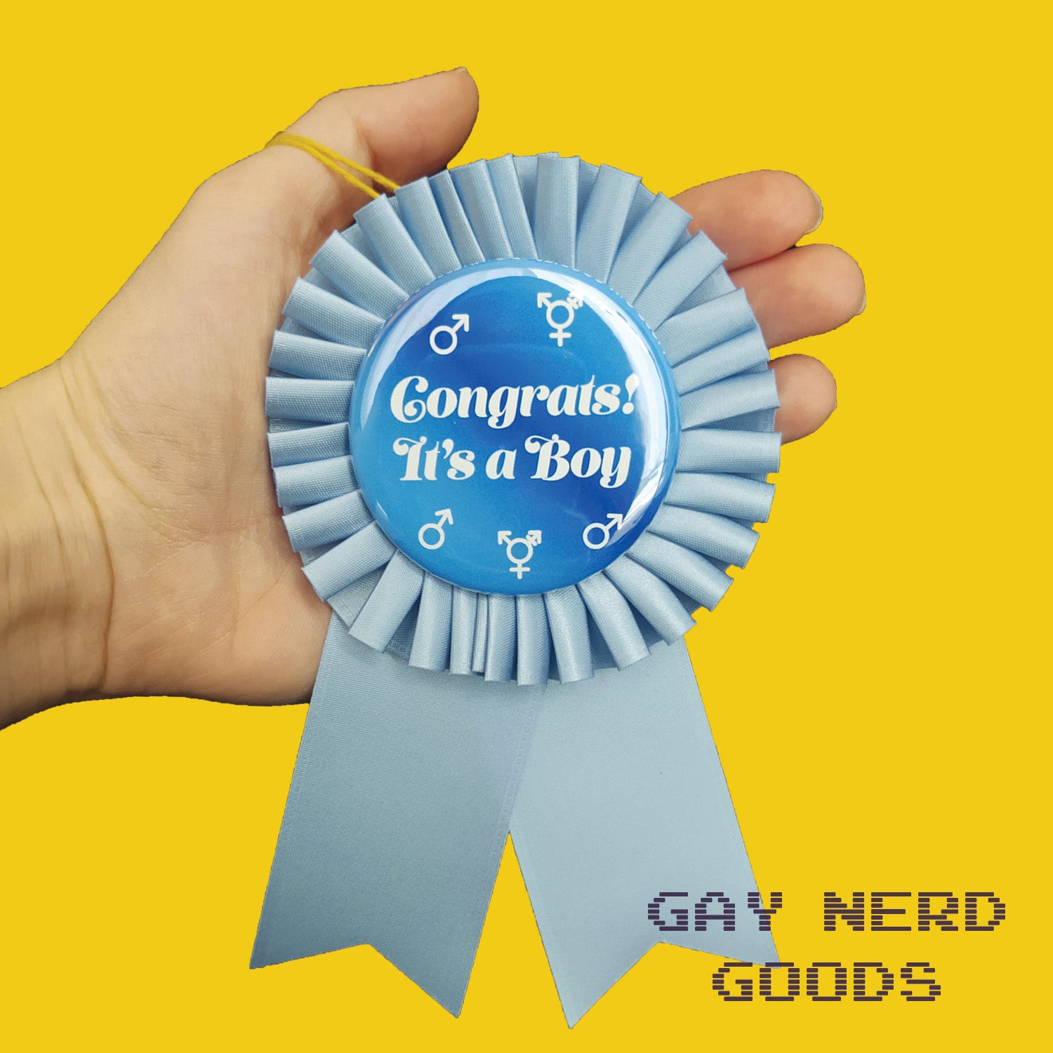 hand holding the blue "congrats! it's a boy" award ribbon rosette on a yellow background