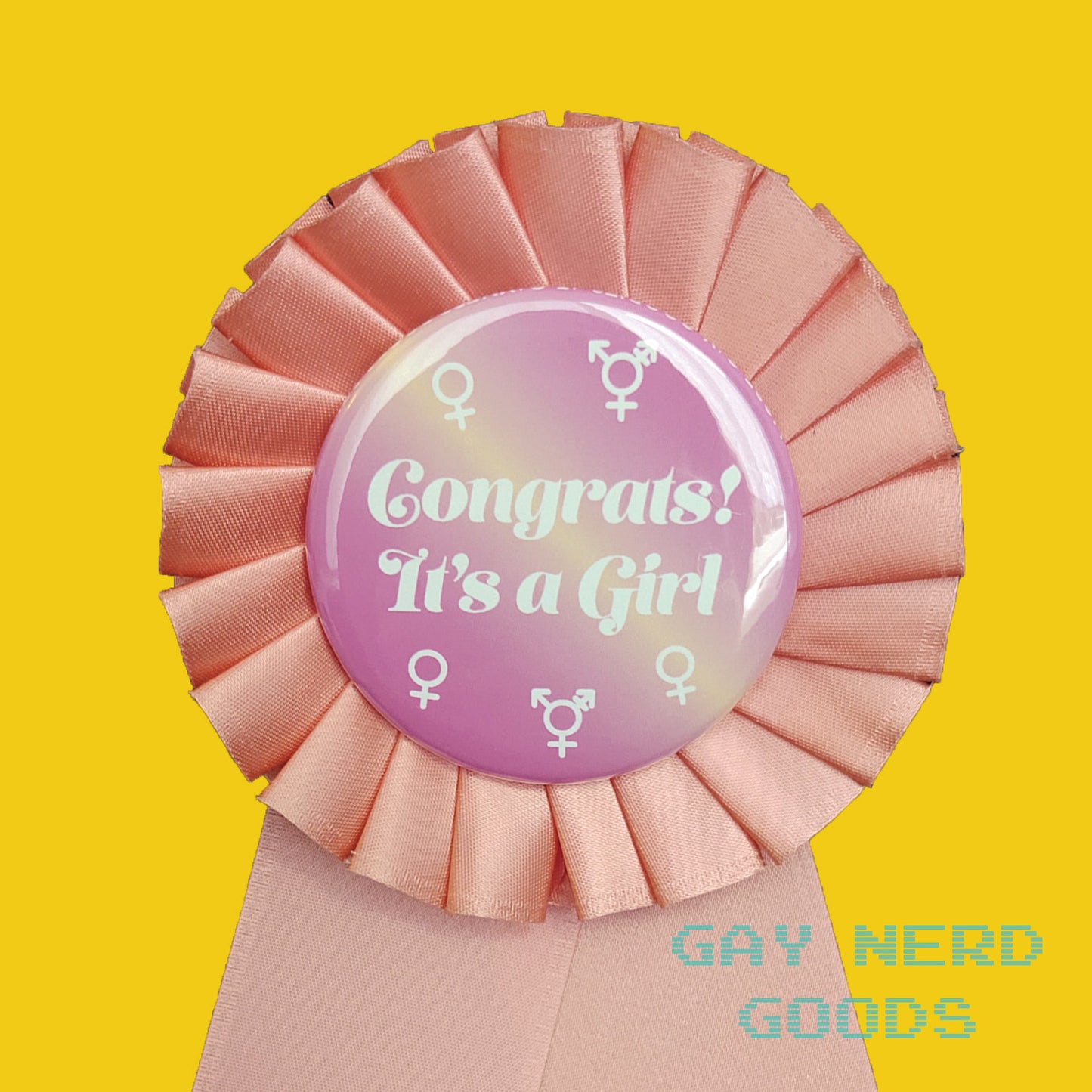 close up detail of the "congrats! it's a girl" award ribbon. The central button has white text and white female and trans symbols on a pink and yellow gradient background. The satin ribbon around it is light pink