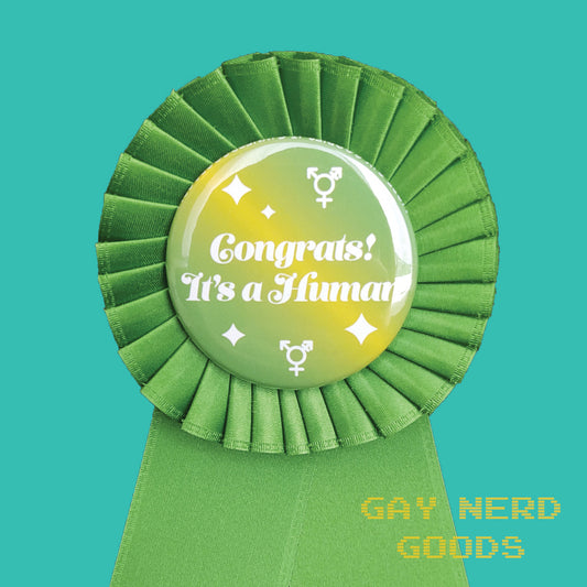 close up view of the "congrats! it's a human" award ribbon. The text is white surrounded by white trans symbols and sparkles. The background of the design is yellow green gradient and the ribbon is green