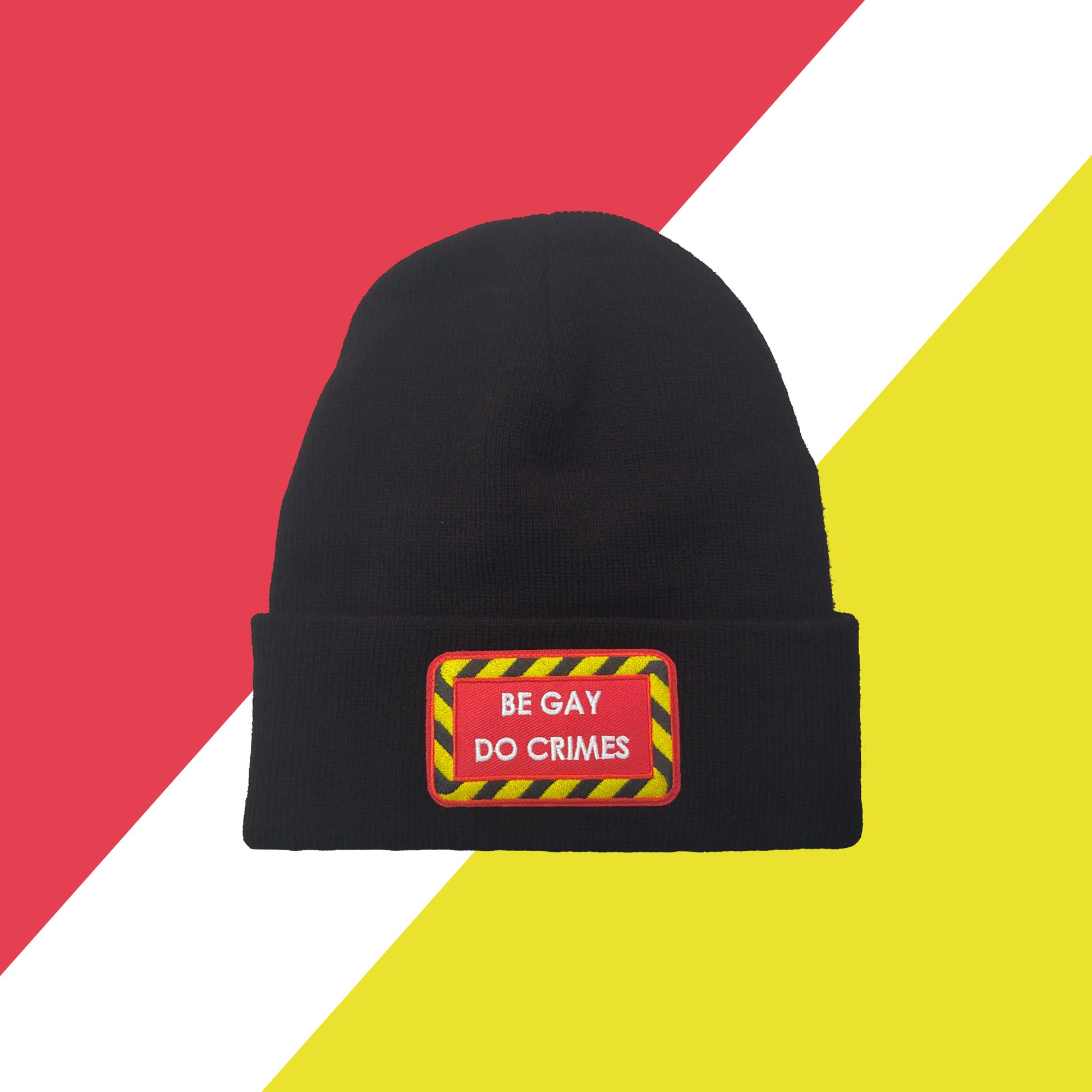 black beanie with red patch on the front with yellow and black striped border and white text reading "be gay do crimes". Background is red white and yellow shapes