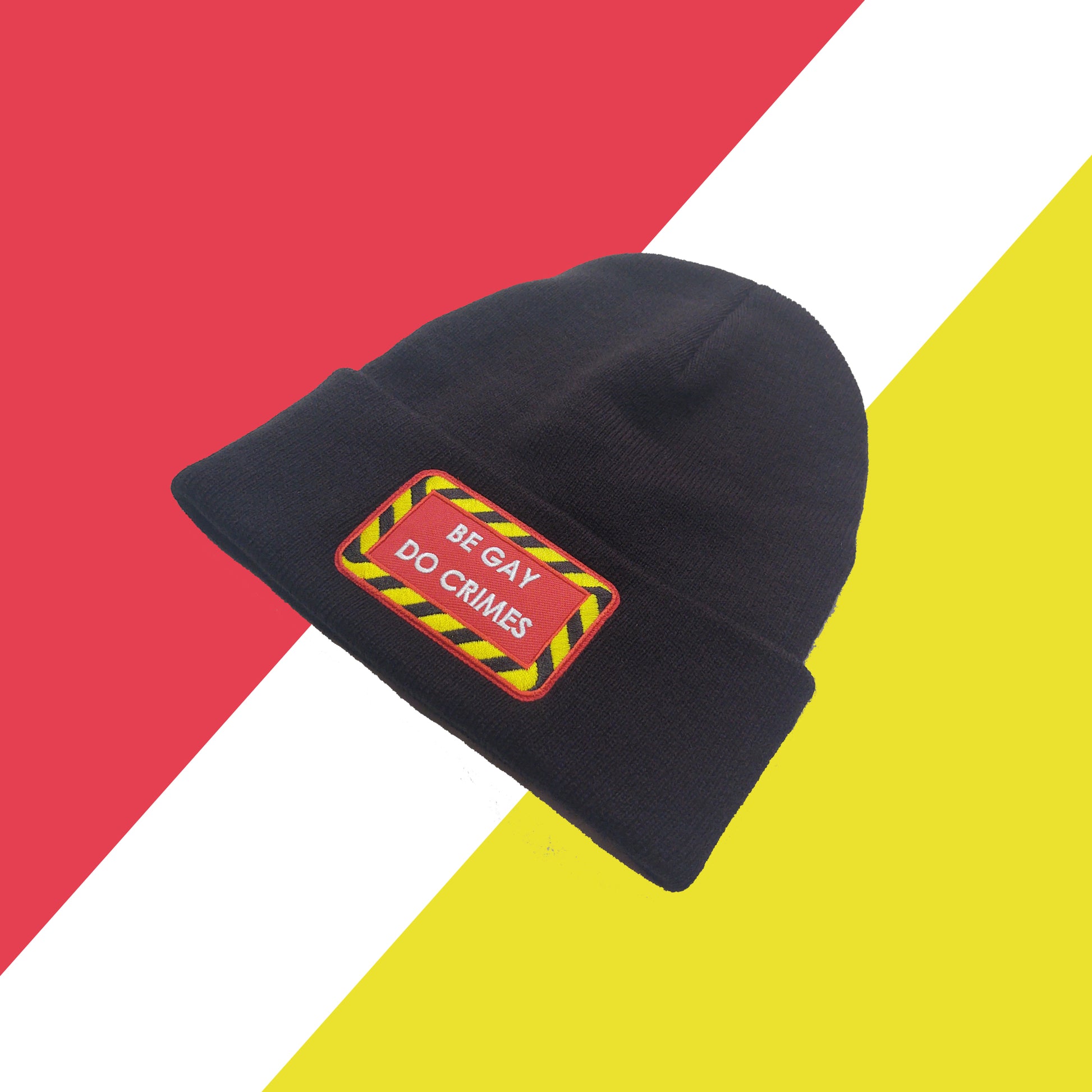black beanie set at an angle with red patch on the front with yellow and black striped border and white text reading "be gay do crimes". Background is red white and yellow shapes