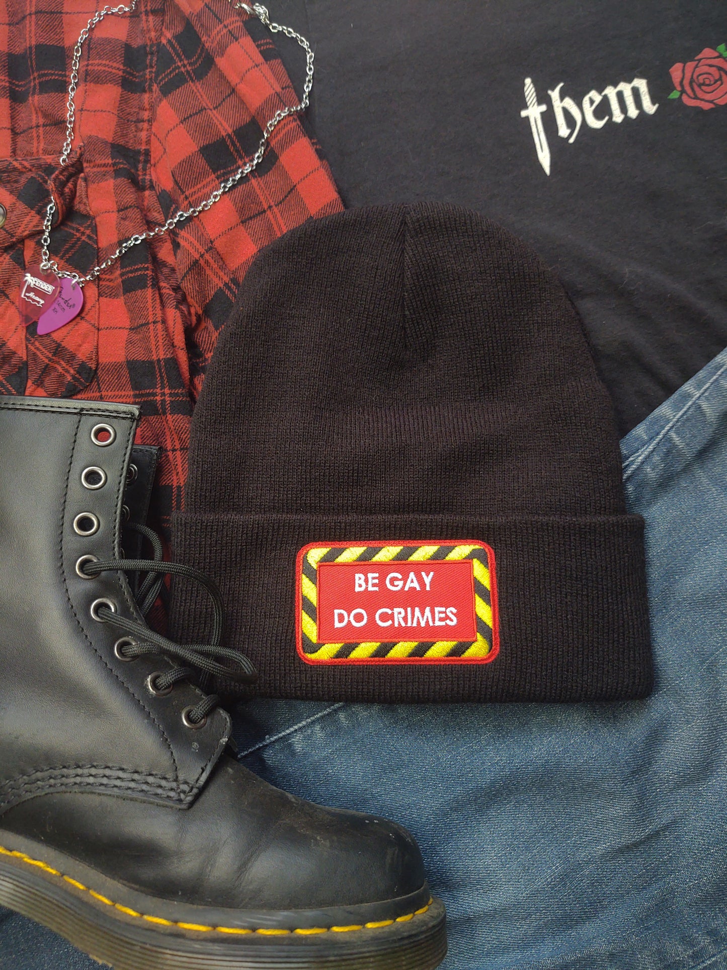 black beanie with red patch on the front with yellow and black striped border and white text reading "be gay do crimes". It is surrounded by the "them fatale" shirt, jeans, a red and black flannel, guitar pick necklace, and black doc martin boots.
