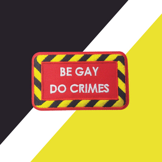 red iron-on patch with a black and yellow striped border and white text "be gay do crimes". The background is black, white, and yellow shapes.