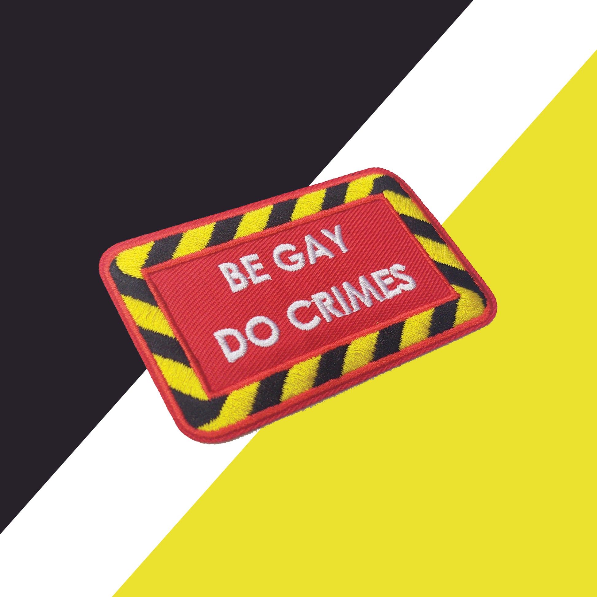 red iron-on patch set at an angle with a black and yellow striped border and white text "be gay do crimes". The background is black, white, and yellow shapes.