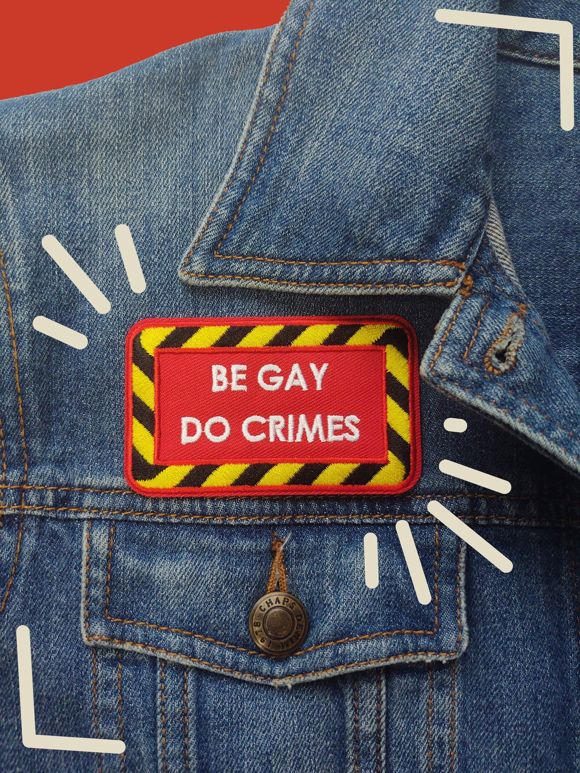 red iron-on patch with a black and yellow striped border and white text "be gay do crimes". It's sitting on the chest of a denim jacket