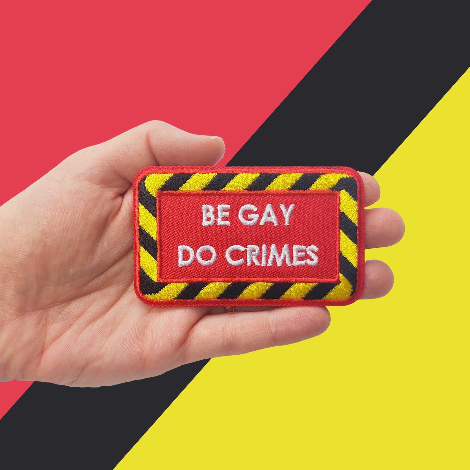 red iron-on patch with a black and yellow striped border and white text "be gay do crimes" held by a light-skinned hand. The background is black, red, and yellow shapes.
