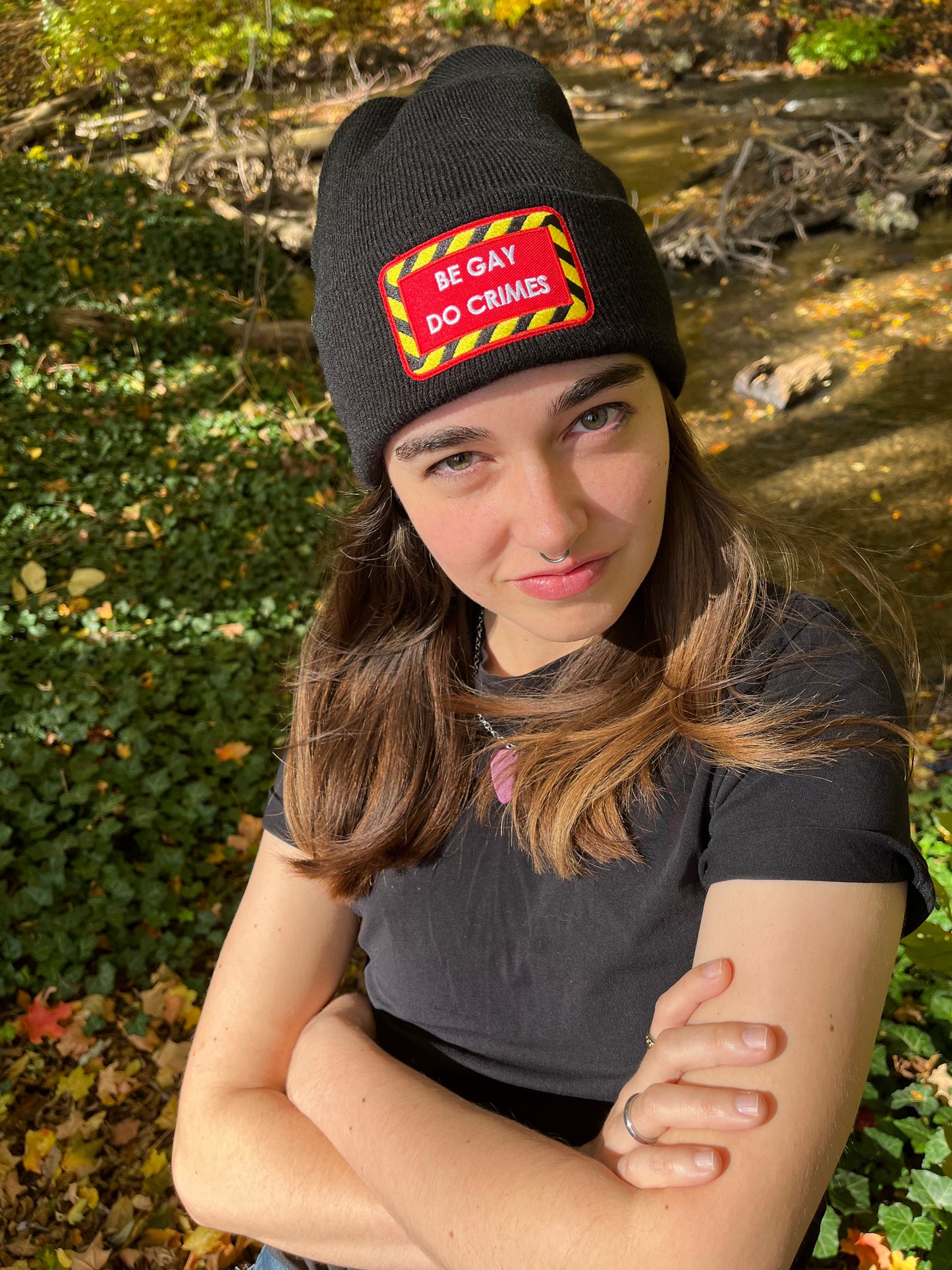 person with arms crossed wearing black "be gay do crimes" beanie against a forest background