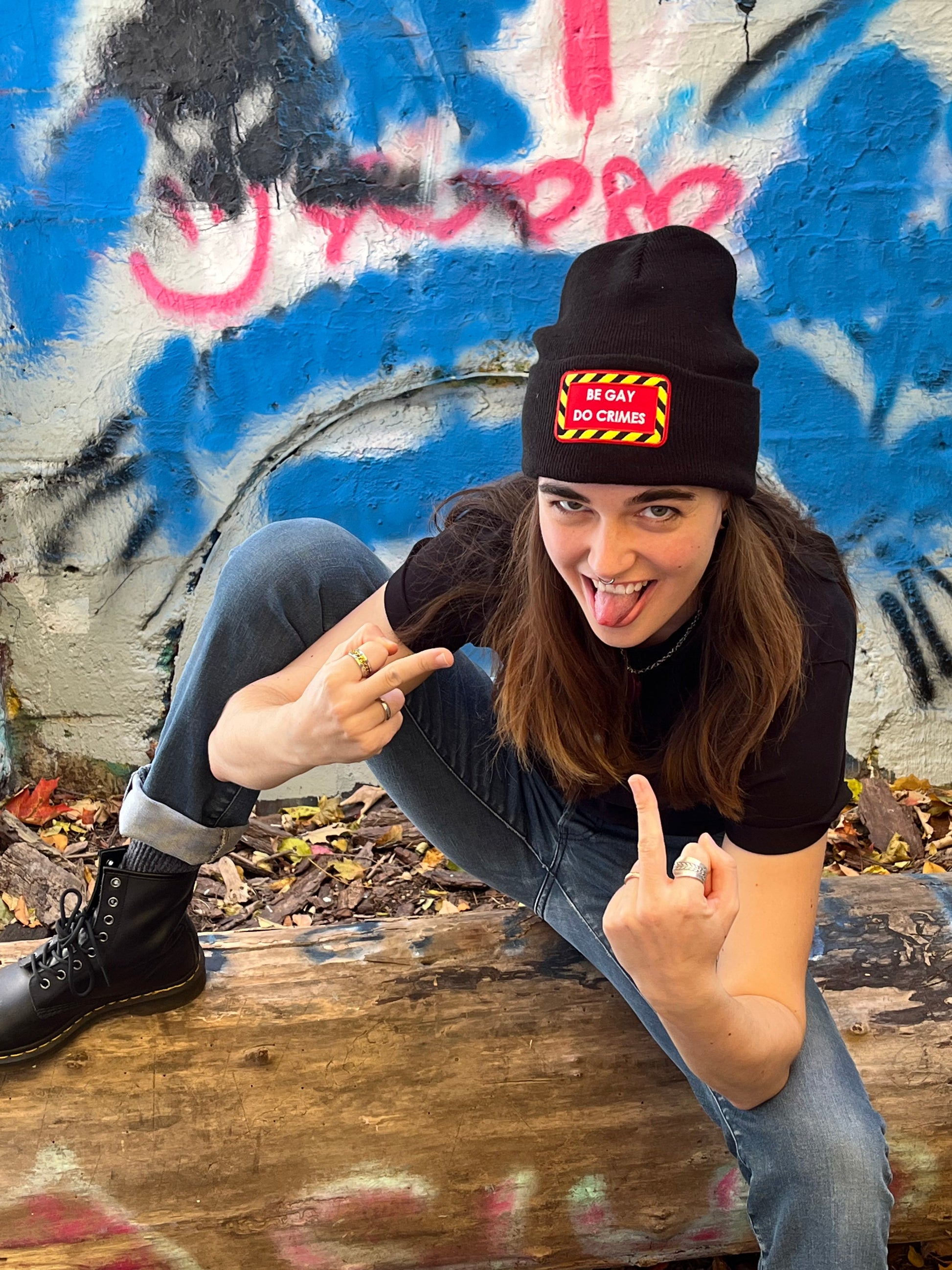 person squatting on a log in front of graffiti flipping off the camera with tongue sticking out wearing the black "be gay do crimes" beanie