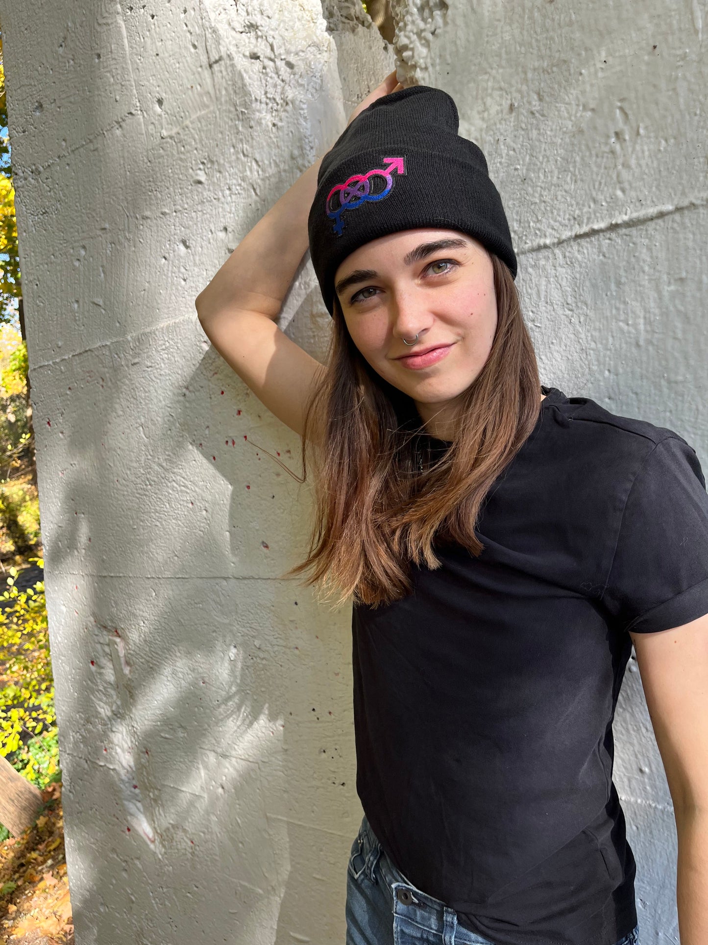 person with their arm behind their head smiling leaning against a white concrete wall wearing the bisexual pride beanie