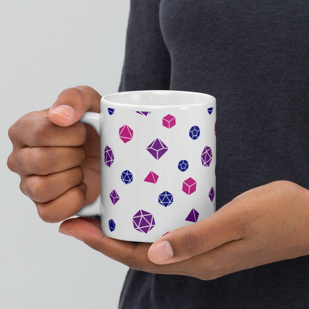 white mug being held by the handle by a dark-skinned set of hands. It has an all-over print of polyhedral d&d dice in the bisexual colors of blue, pink, and purple