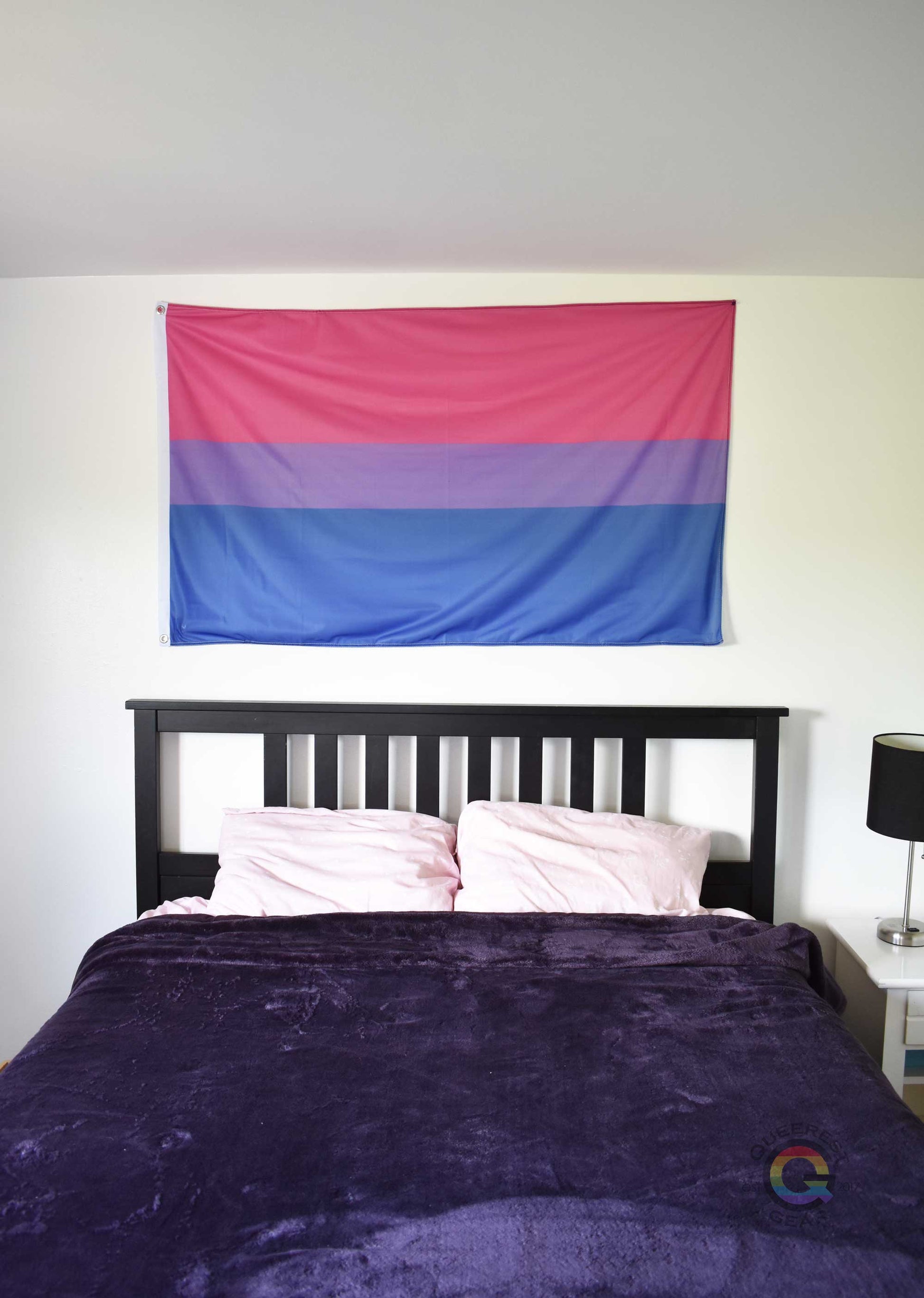3’x5’ bisexual pride flag hanging horizontally on the wall of a bedroom centered above a bed with a purple blanket