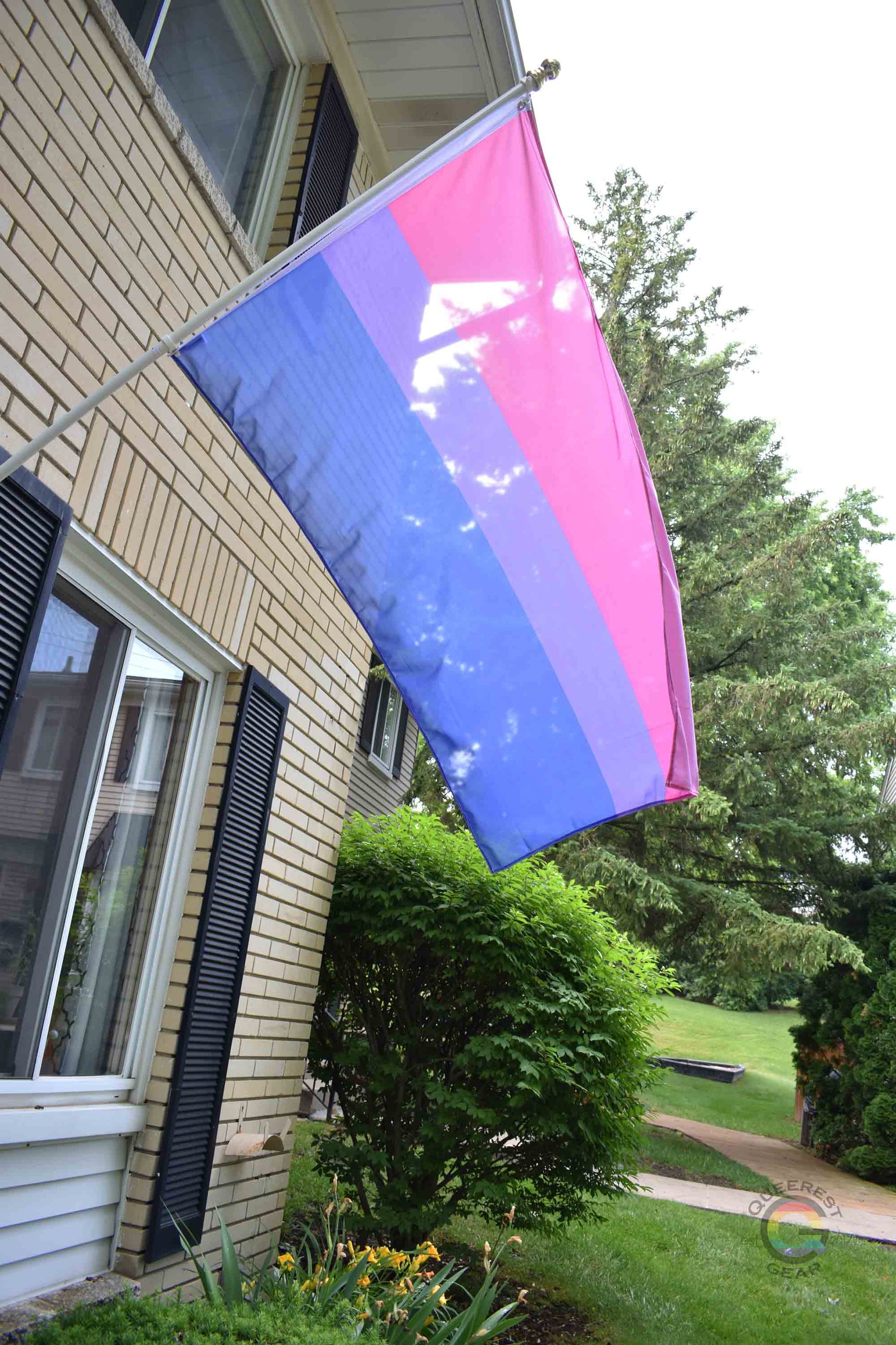3’x5’ bisexual pride flag hanging from a flagpole on the outside of a light brick house with dark shutters