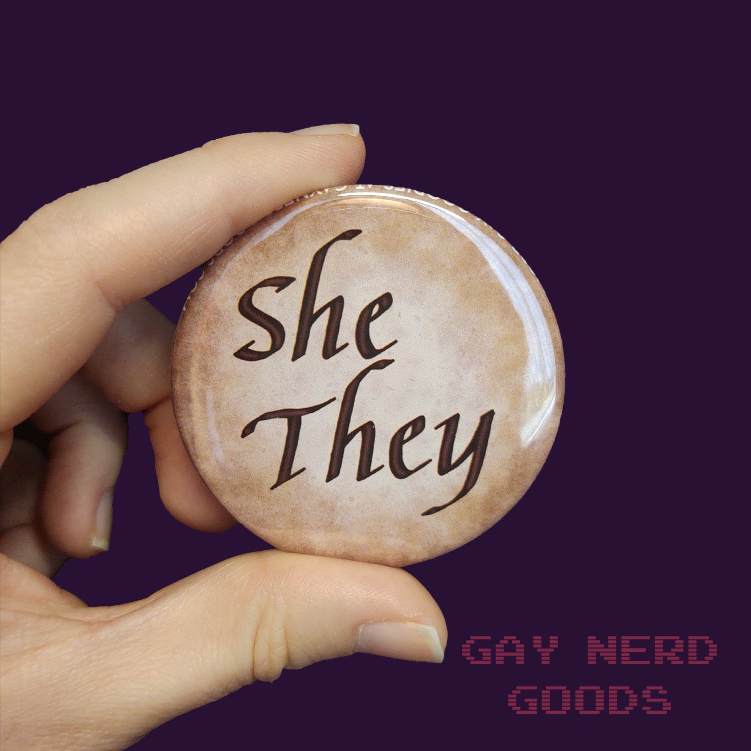 she they large calligraphy pronoun button held in two fingers on a dark purple background