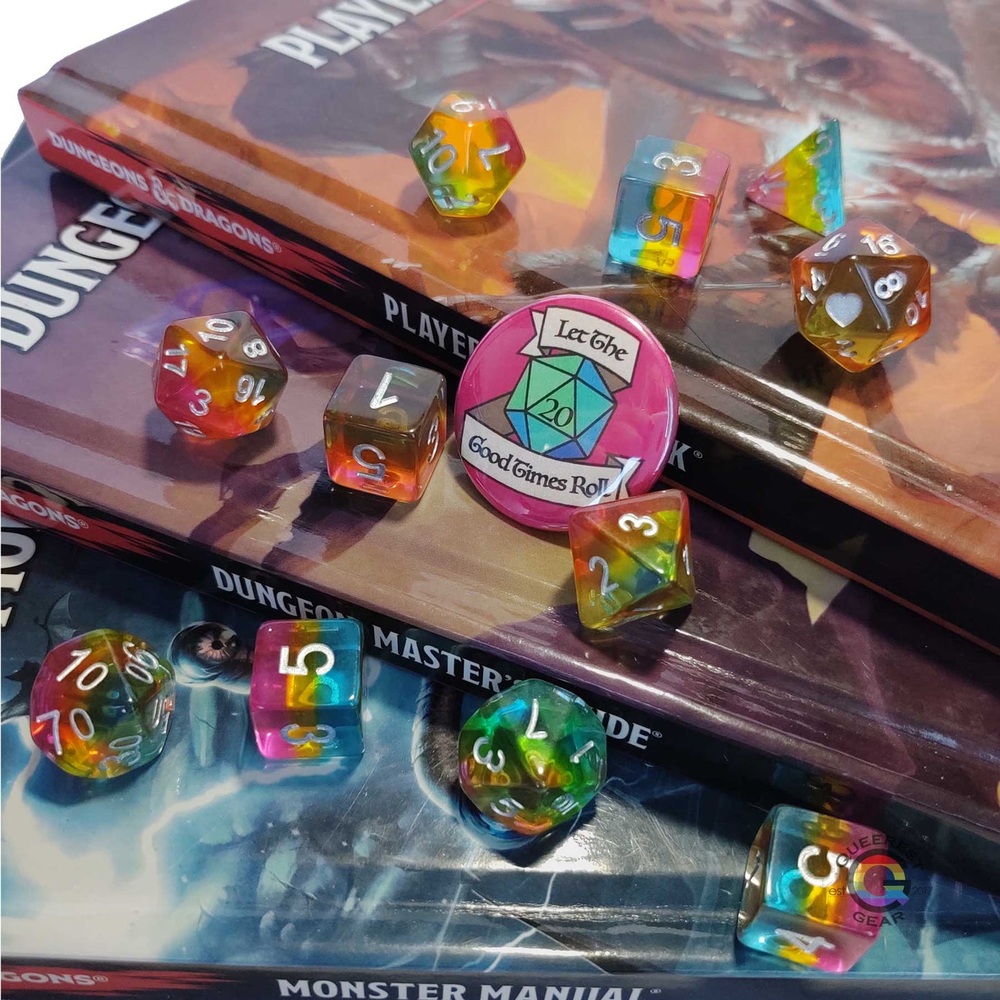 11 piece set of pansexual dice scattered on a stack of D&D books. They are transparent and colored in the stripes of the nonbinary flag with silver ink. There is the freebie “let the good times roll” pinback button among them. 