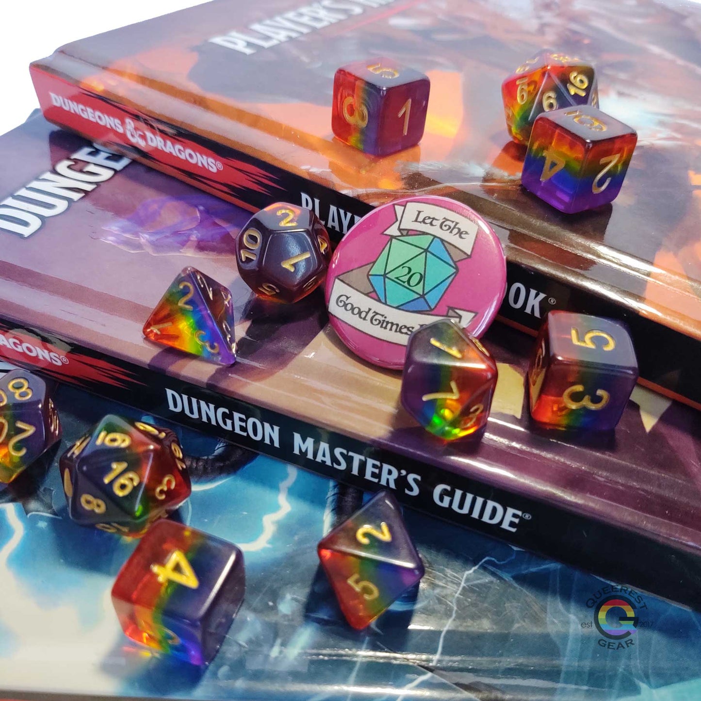 11 piece set of polyhedral dice scattered on a stack of D&D books. They are transparent and colored in the stripes of the rainbow flag with gold ink. There is the freebie “let the good times roll” pinback button among them. 