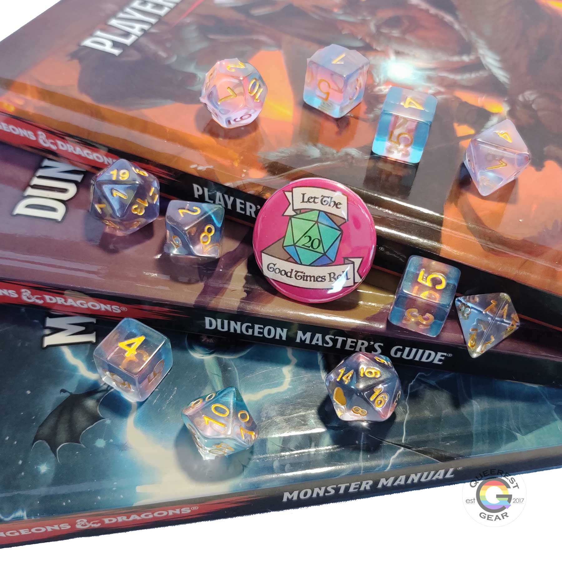 11 piece set of polyhedral dice scattered on a stack of D&D books. They are transparent and colored in the stripes of the transgender flag with gold ink. There is the freebie “let the good times roll” pinback button among them.