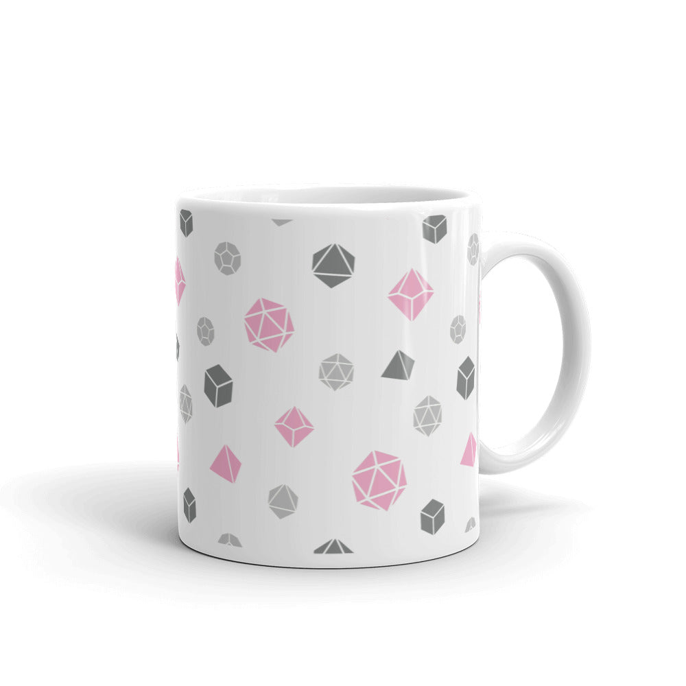 white mug on a white background with handle facing right. It has an all-over print of polyhedral d&d dice in the demigirl colors of light grey, dark grey, and pink