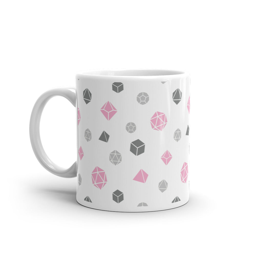 white mug on a white background with handle facing left. It has an all-over print of polyhedral d&d dice in the demigirl colors of dark grey, light grey, and pink