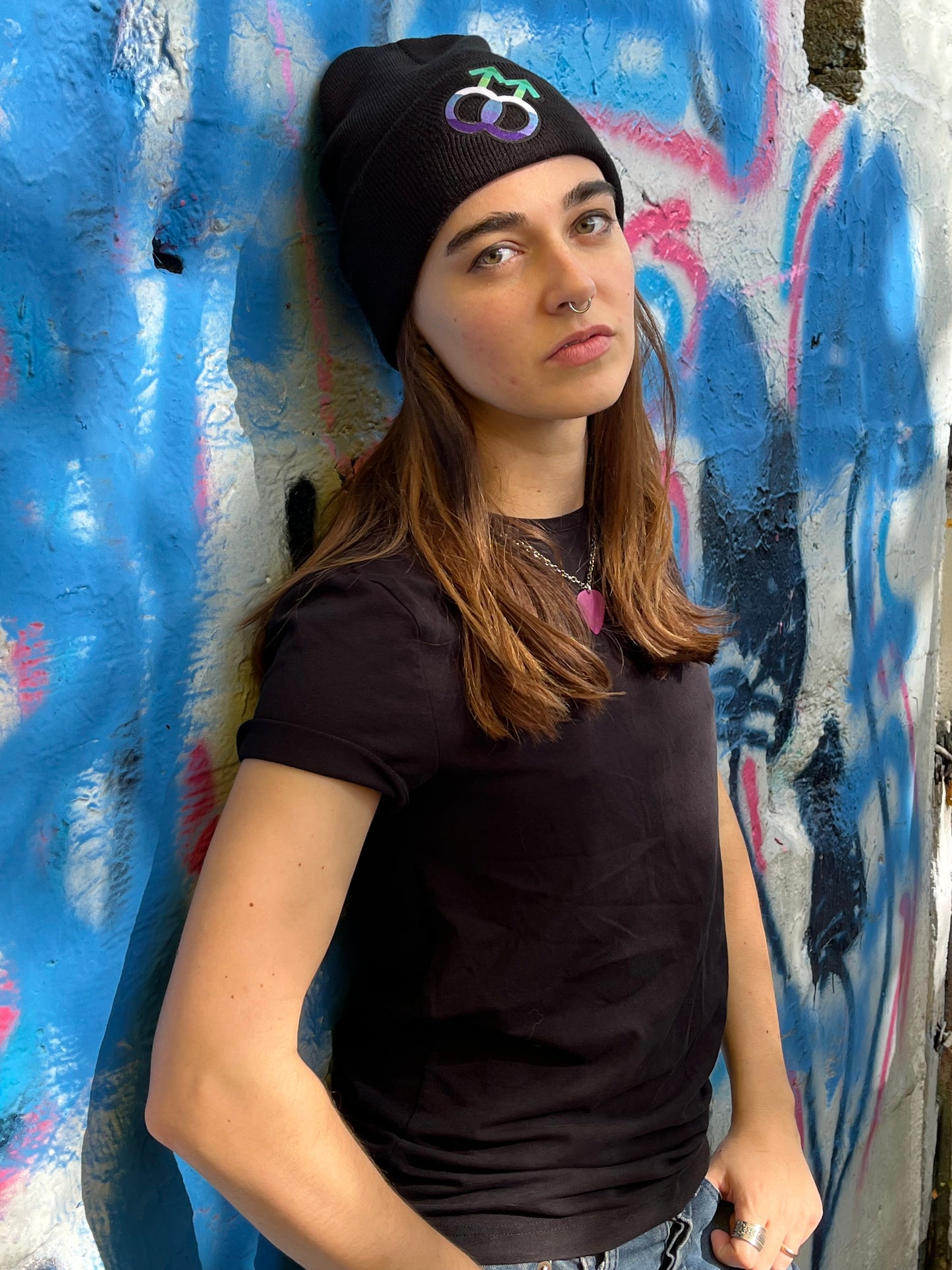 waist up shot of person leaning against graffitied wall wearing the gay mlm beanie