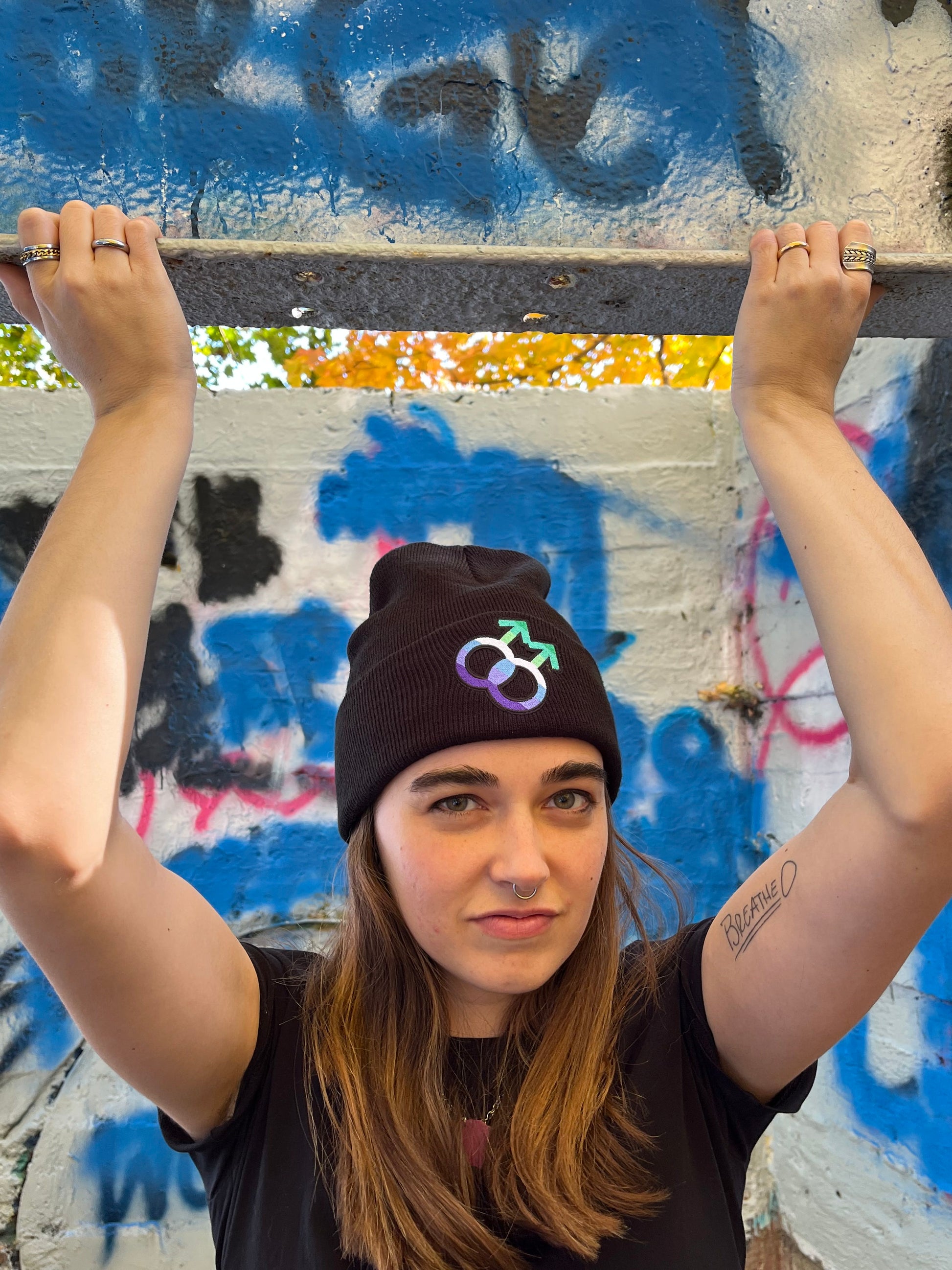 chest up shot of person with arms above head gripping an I-beam. They are wearing the black gay mlm symbol beanie