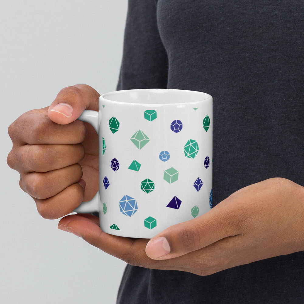 white mug being held by the handle by a dark-skinned set of hands. It has an all-over print of polyhedral d&d dice in the gay mlm colors of greens and blues