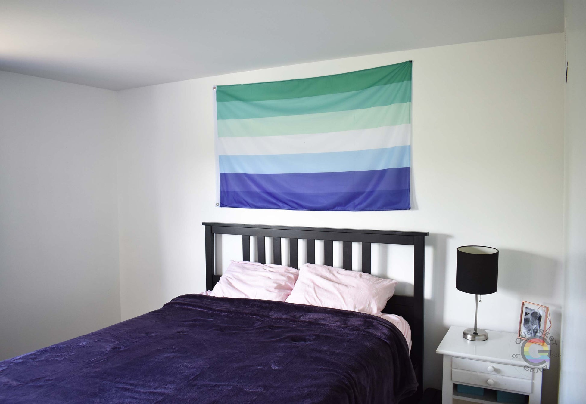 3’x5’ gay mlm pride flag hanging horizontally on the wall of a bedroom centered above a bed with a purple blanket