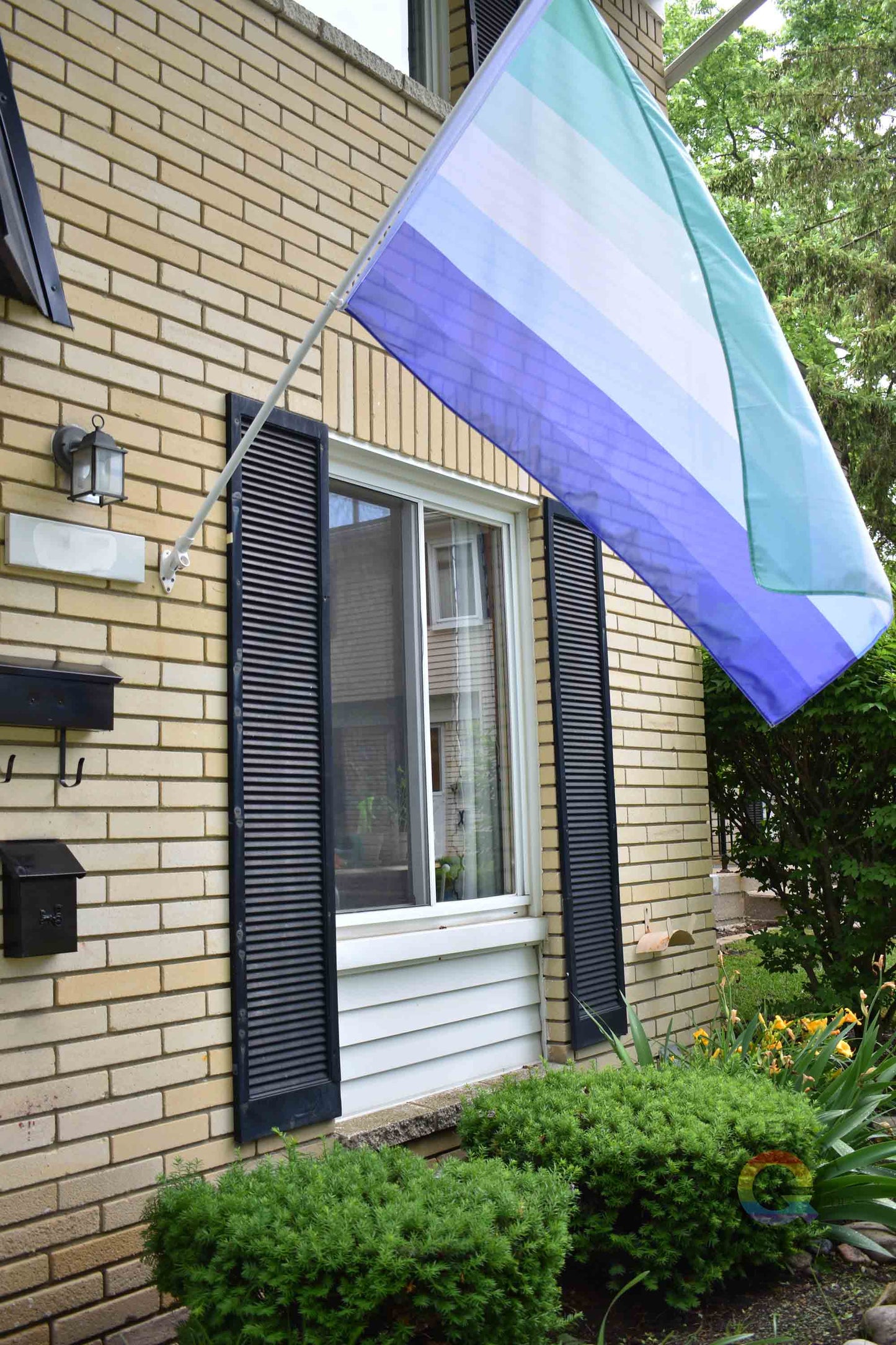 3’x5’ gay pride flag hanging from a flagpole on the outside of a light brick house with dark shutters