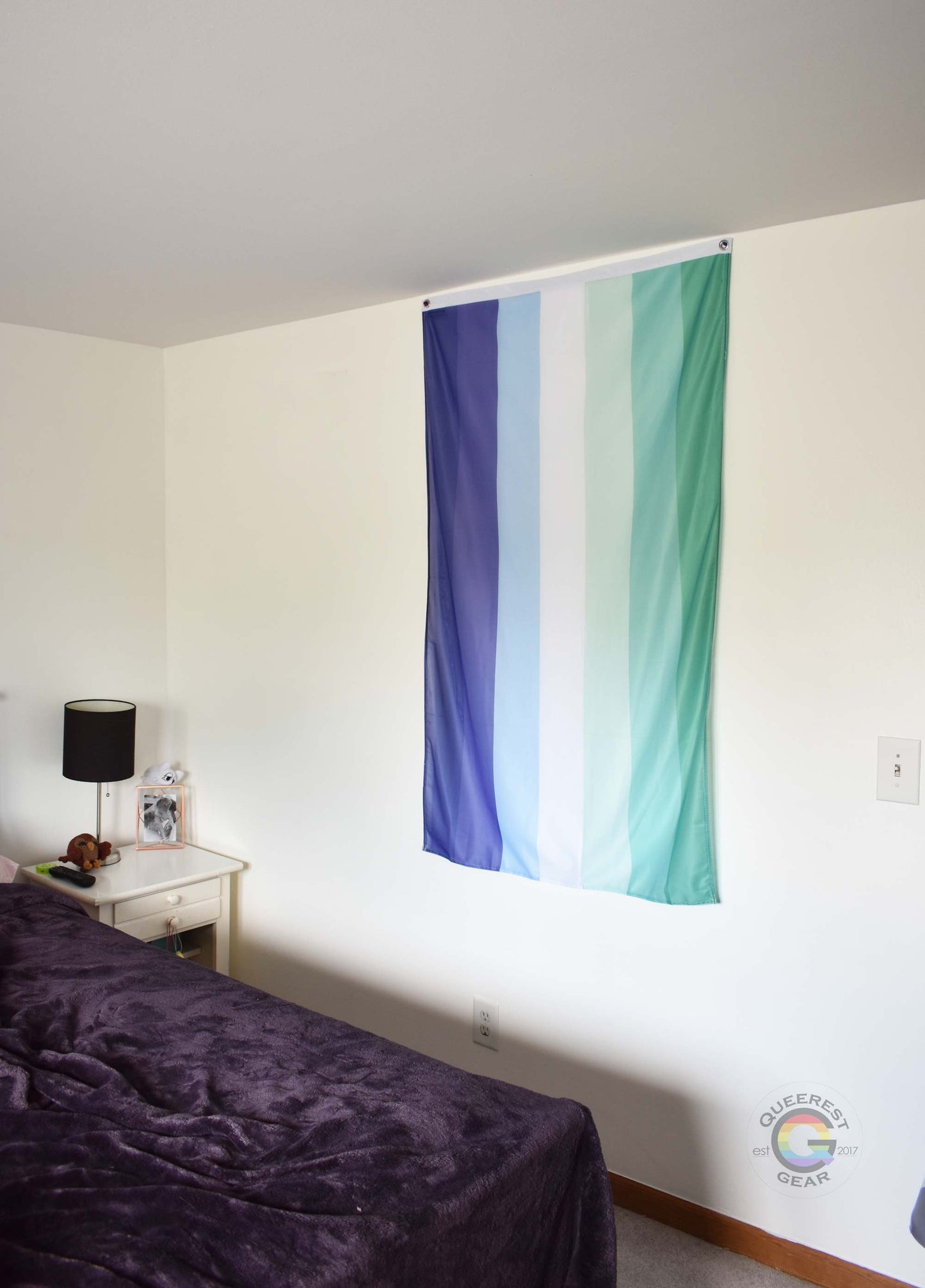  3’x5’ gay pride flag hanging vertically on the wall of a bedroom with a nightstand and a bed