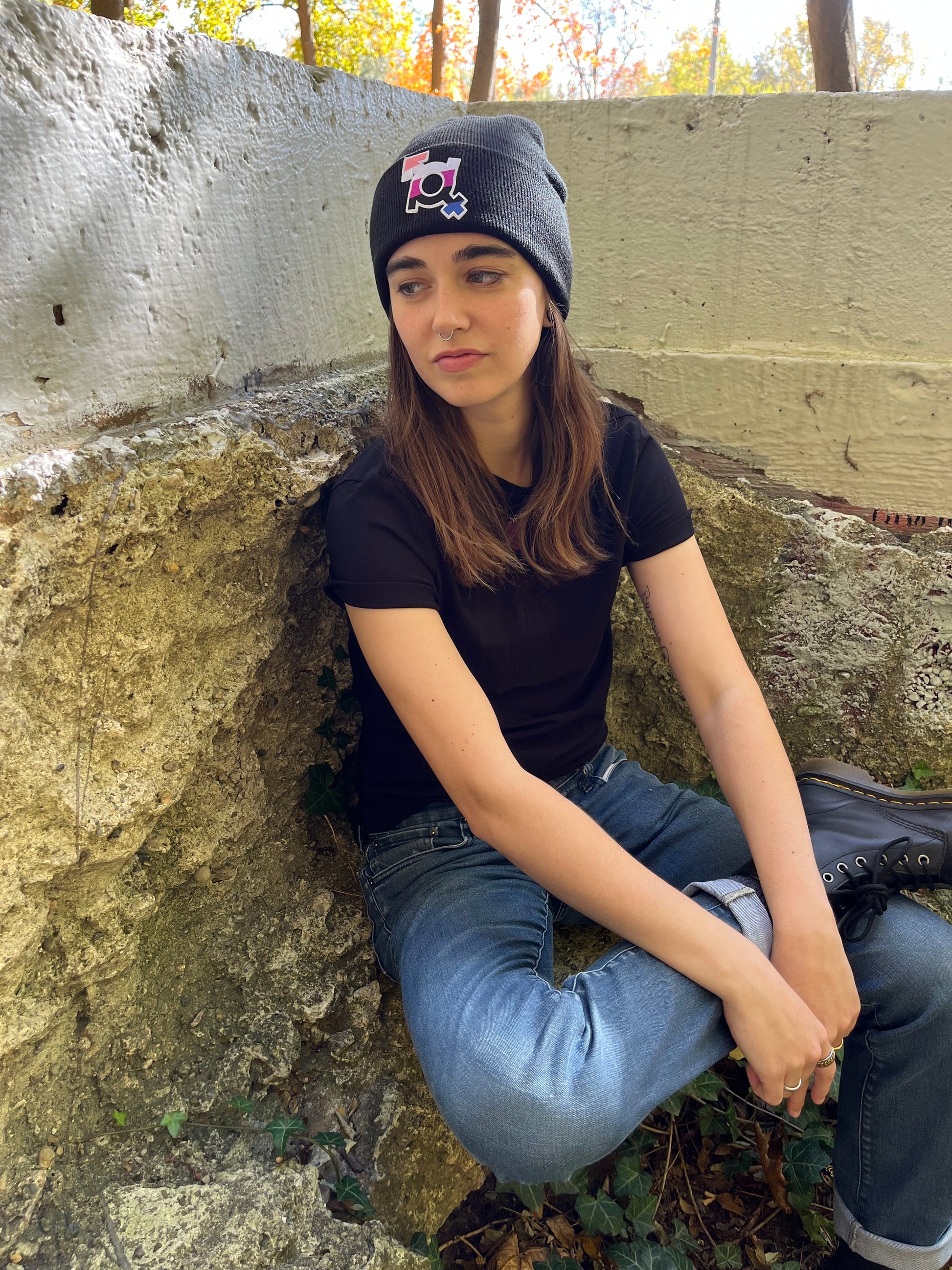 person sitting with legs crossed on concreted slab looking to the side and wearing the genderfluid beanie