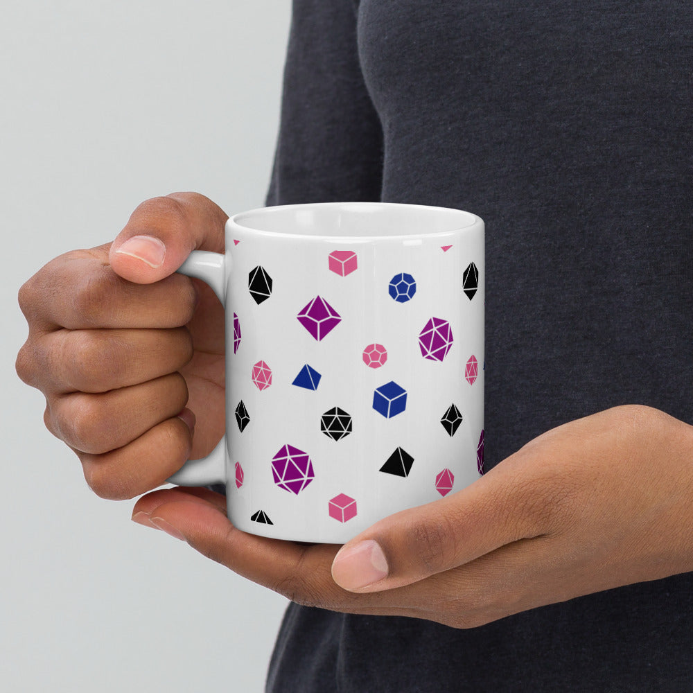 white mug being held by the handle by a dark-skinned set of hands. It has an all-over print of polyhedral d&d dice in the genderfluid colors of peach, magenta, black, and blue