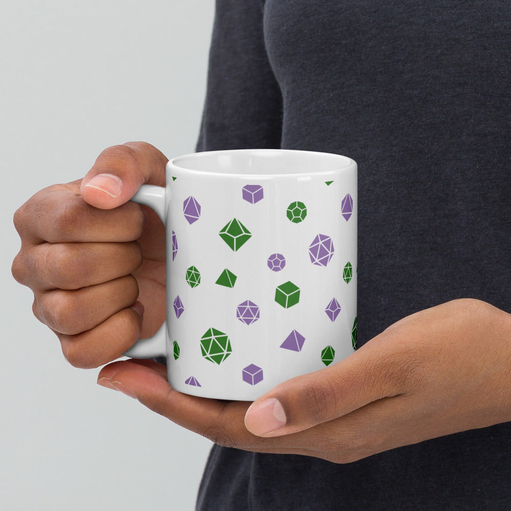 white mug being held by the handle by a dark-skinned set of hands. It has an all-over print of polyhedral d&d dice in the genderqueer colors of green and purple