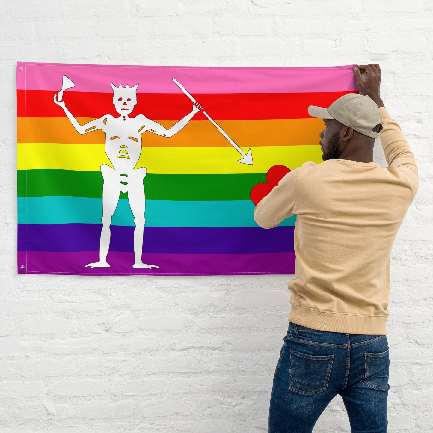 the gilbert baker flag with stripes of pink, red, orange, yellow, green, turquoise, indigo, and purple and blackbeard's symbol being hung on the wall by a person with dark skin and a beard