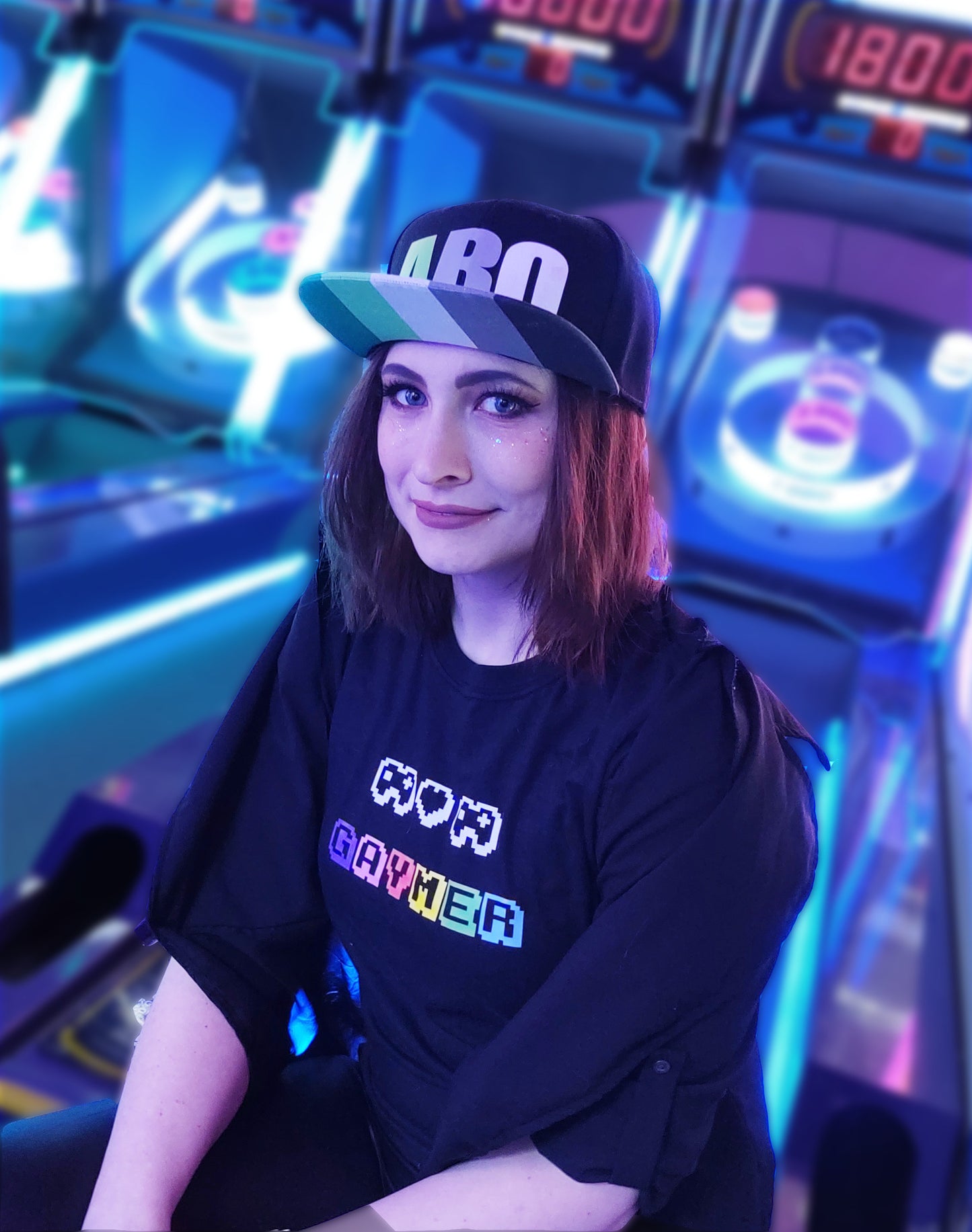 A model wears the aromantic pride hat. They are sitting on a skeeball machine and wearing a black “gaymer” t-shirt