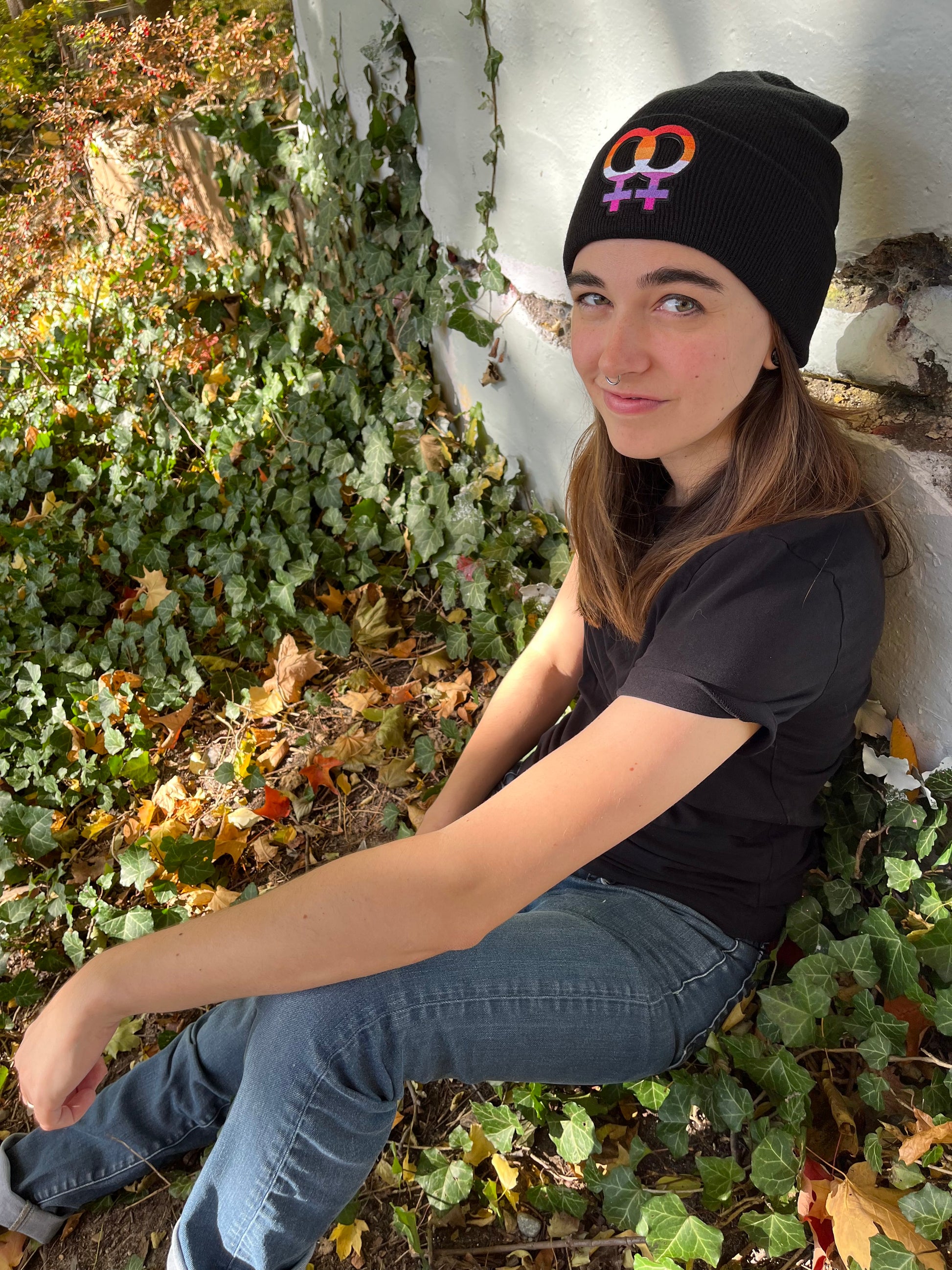 person from the side sitting and leaning against an ivy covered wall wearing the lesbian pride beanie