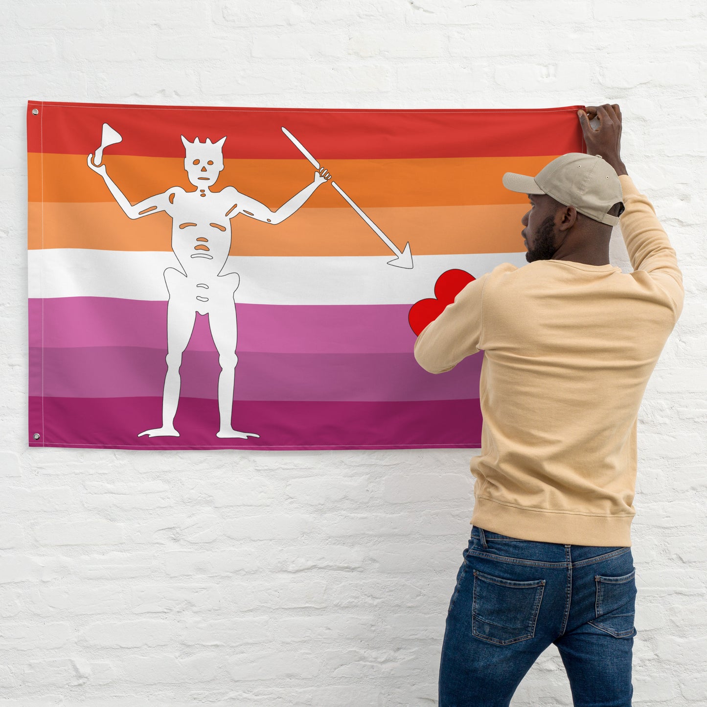 person with dark skin and beard hanging the lesbian blackbeard pirate flag on a white brick wall