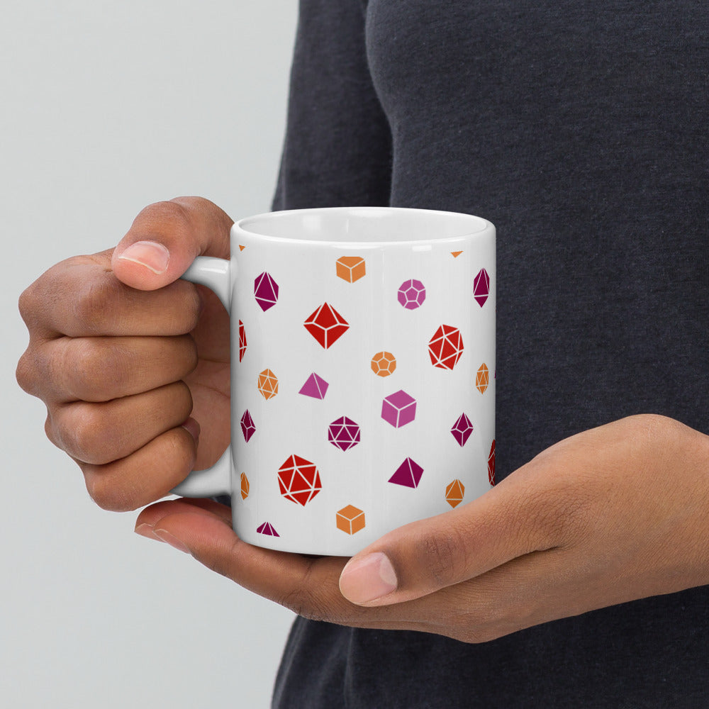 white mug being held by the handle by a dark-skinned set of hands. It has an all-over print of polyhedral d&d dice in the lesbian colors of pinks and oranges