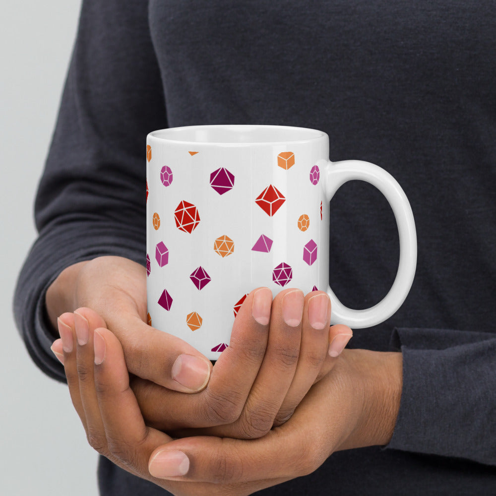white mug being cupped by a dark-skinned set of hands. It has an all-over print of polyhedral d&d dice in the lesbian colors of pinks and oranges