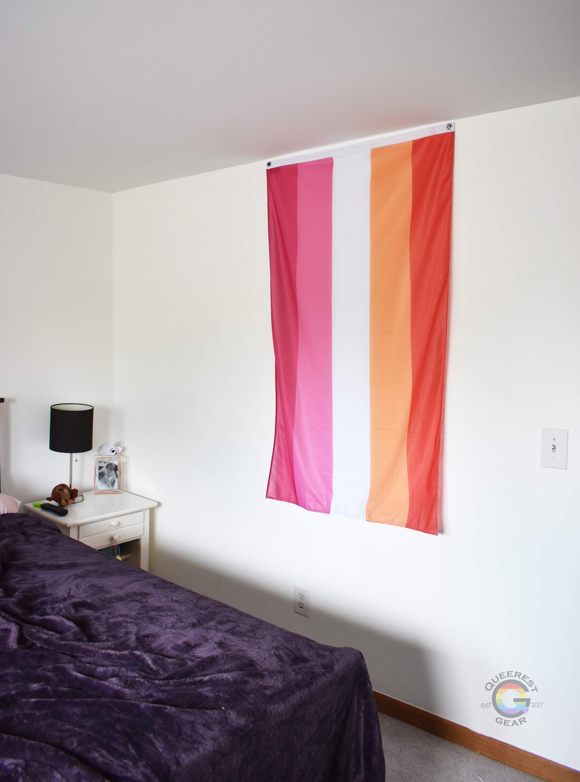  3’x5’ lesbian flag hanging vertically on the wall of a bedroom with a nightstand and a bed