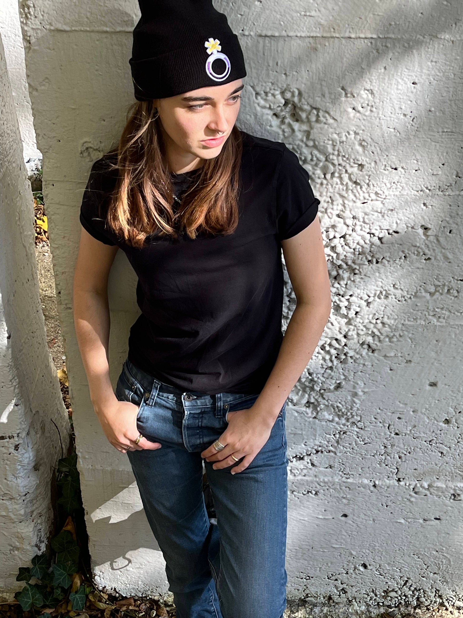 person standing leaning against a white concrete wall with thumbs in pockets wearing a nonbinary beanie
