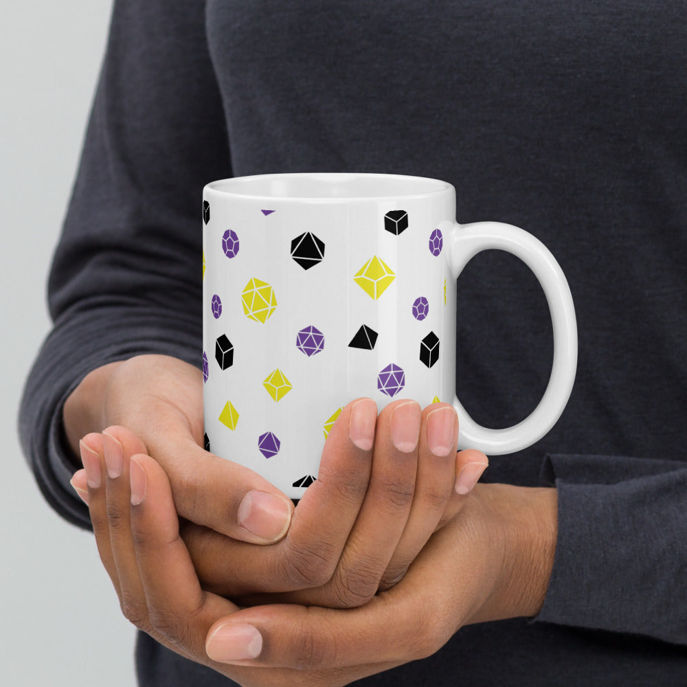 white mug being cupped by a dark-skinned set of hands. It has an all-over print of polyhedral d&d dice in the nonbinary colors of yellow, purple, and black