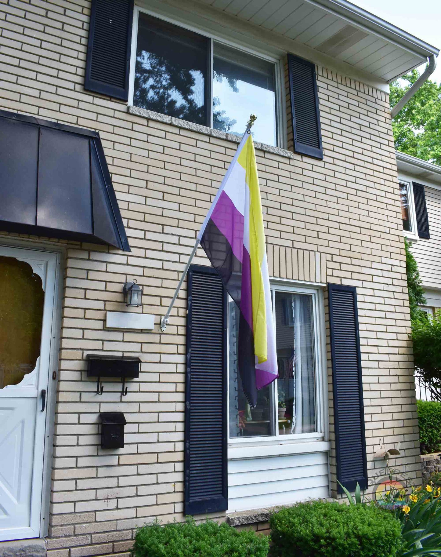 3’x5’ nonbinary pride flag hanging from a flagpole on the outside of a light brick house with dark shutters