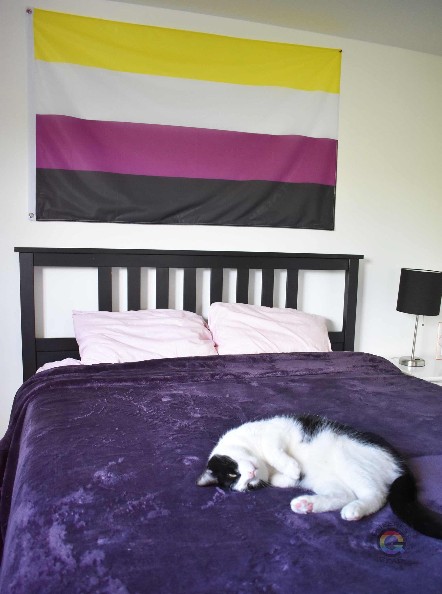3’x5’ nonbinary pride flag hanging horizontally on the wall of a bedroom centered above a bed with a purple blanket. there's a black and white cat laying on the bed