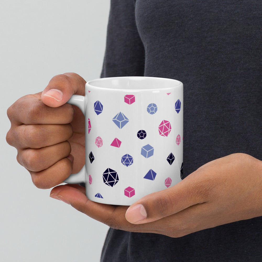 white mug being held by the handle by a dark-skinned set of hands. It has an all-over print of polyhedral d&d dice in the omnisexual colors of pinks, blues, and dark purple