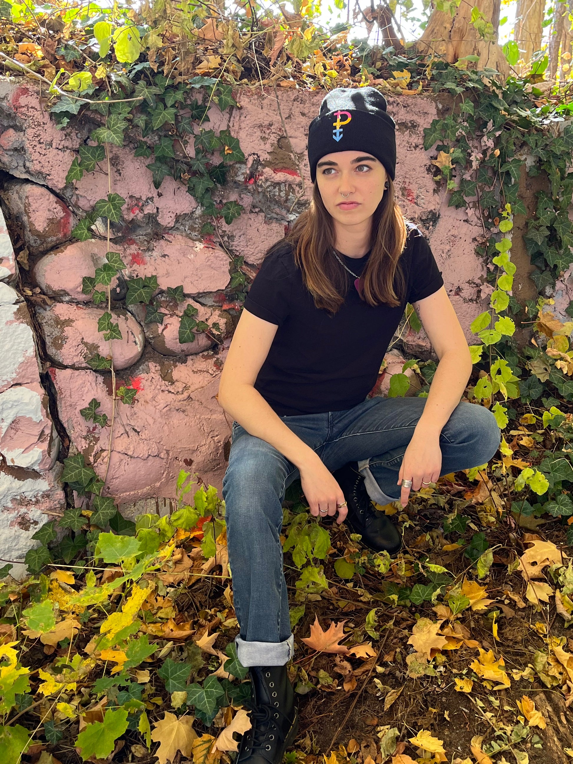 person crouching in front of crumbling ivy covered wall wearing pansexual beanie