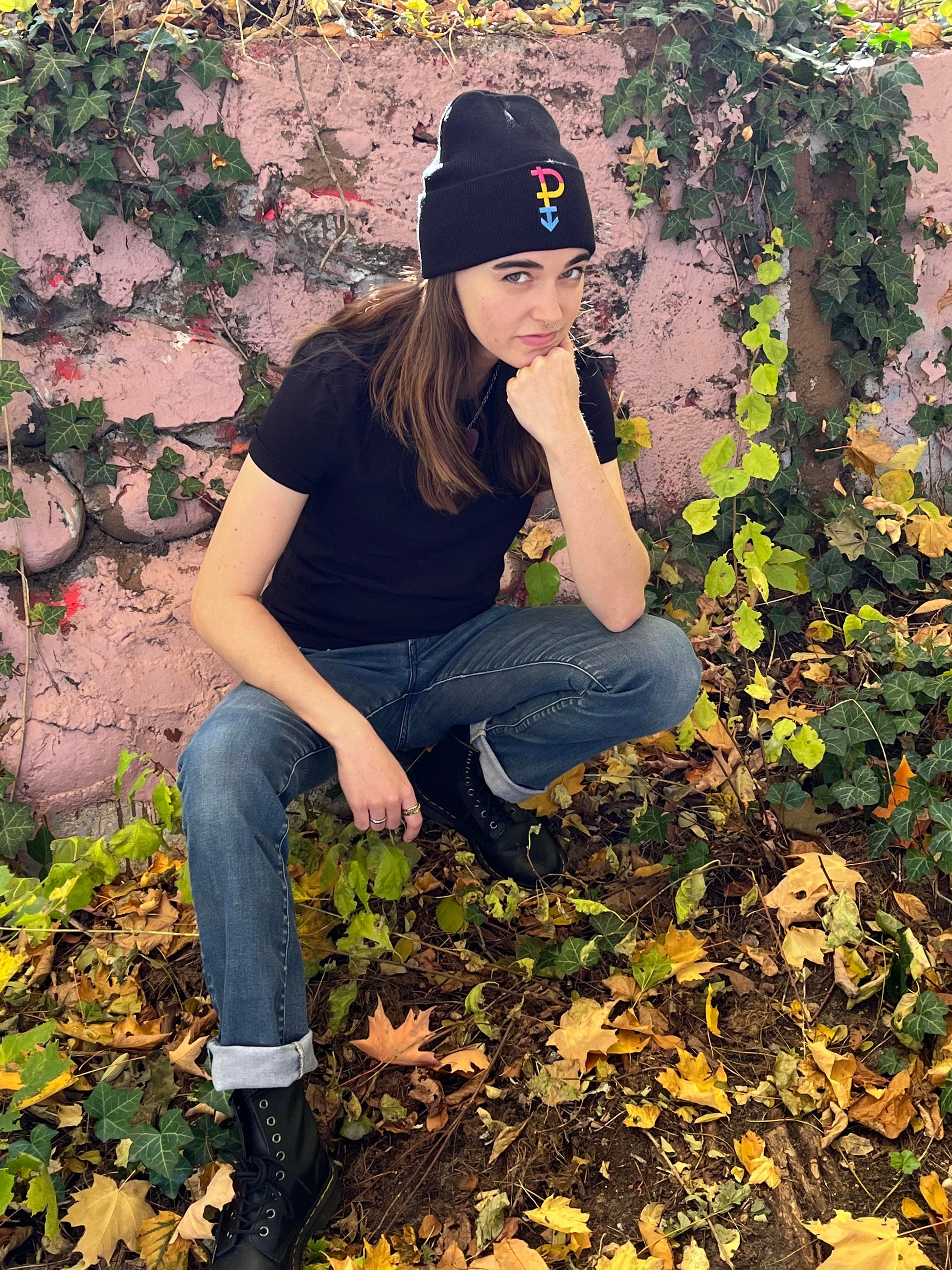 person posing like "the thinker" statue crouching in front of ivy covered wall wearing a pansexual pride beanie